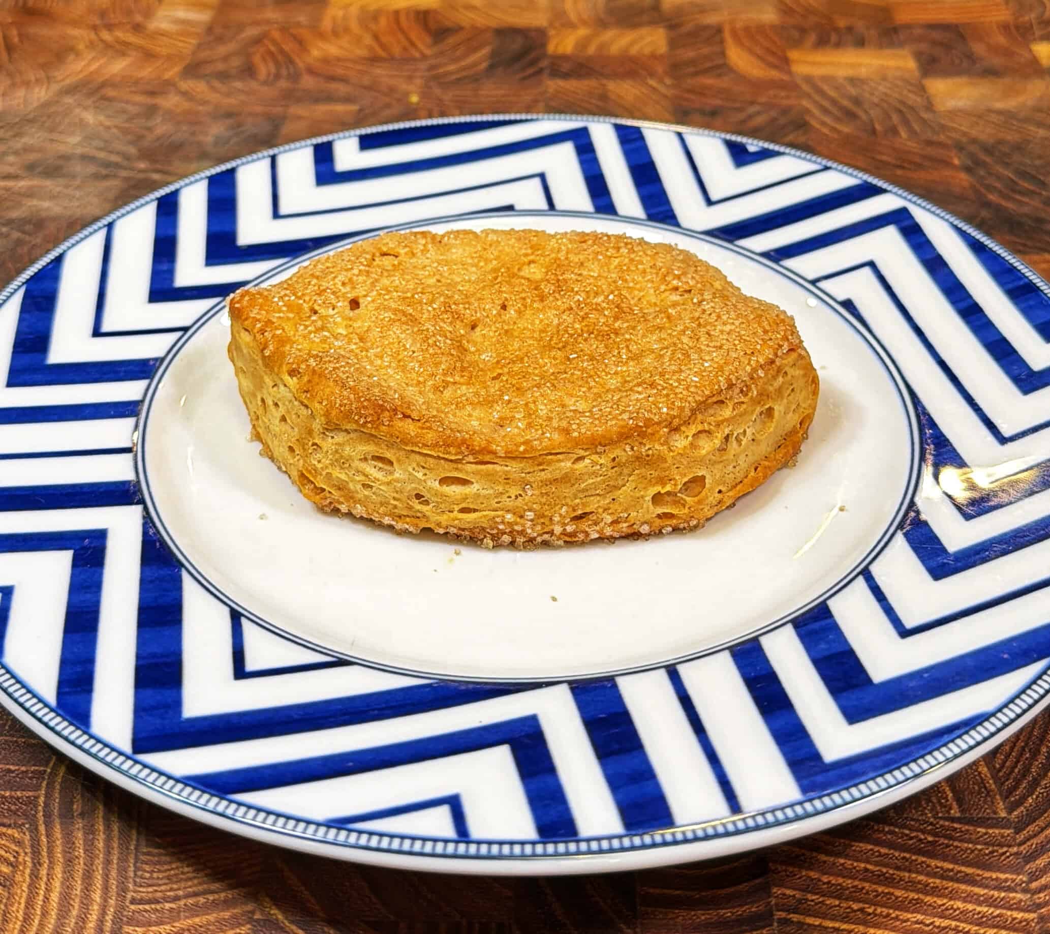 A golden-brown English muffin sits on a white plate with bold blue chevron patterns, atop a wooden surface.