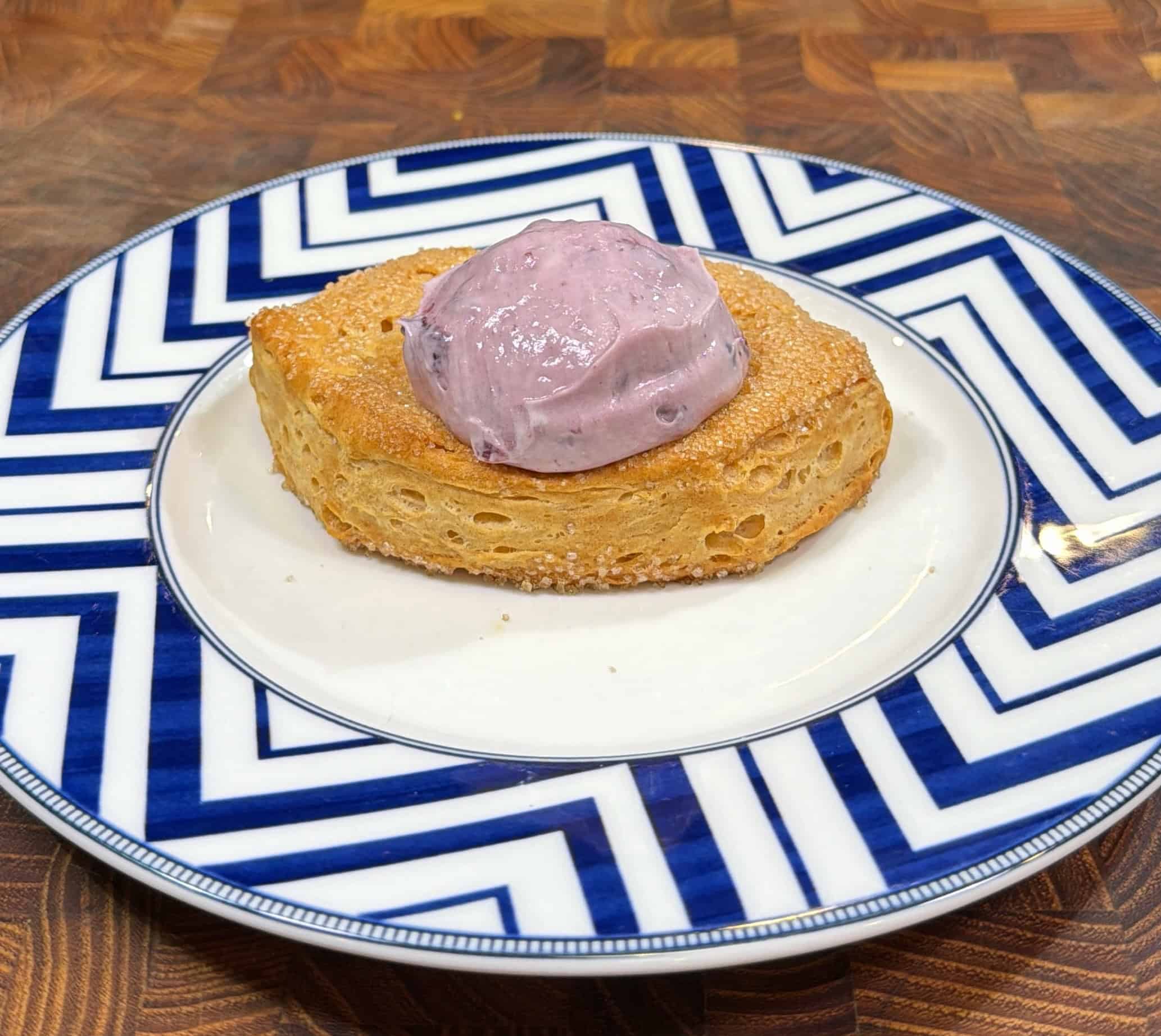 A golden-brown crumpet topped with a dollop of purple cream sits on a white plate with bold blue zigzag patterns, placed on a wooden surface.