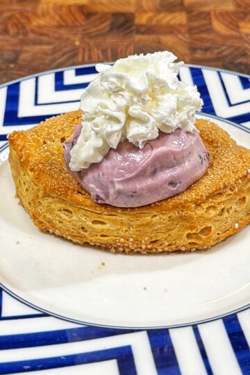 A piece of toasted bread topped with a scoop of purple berry cream and a swirl of whipped cream, served on a blue and white patterned plate.