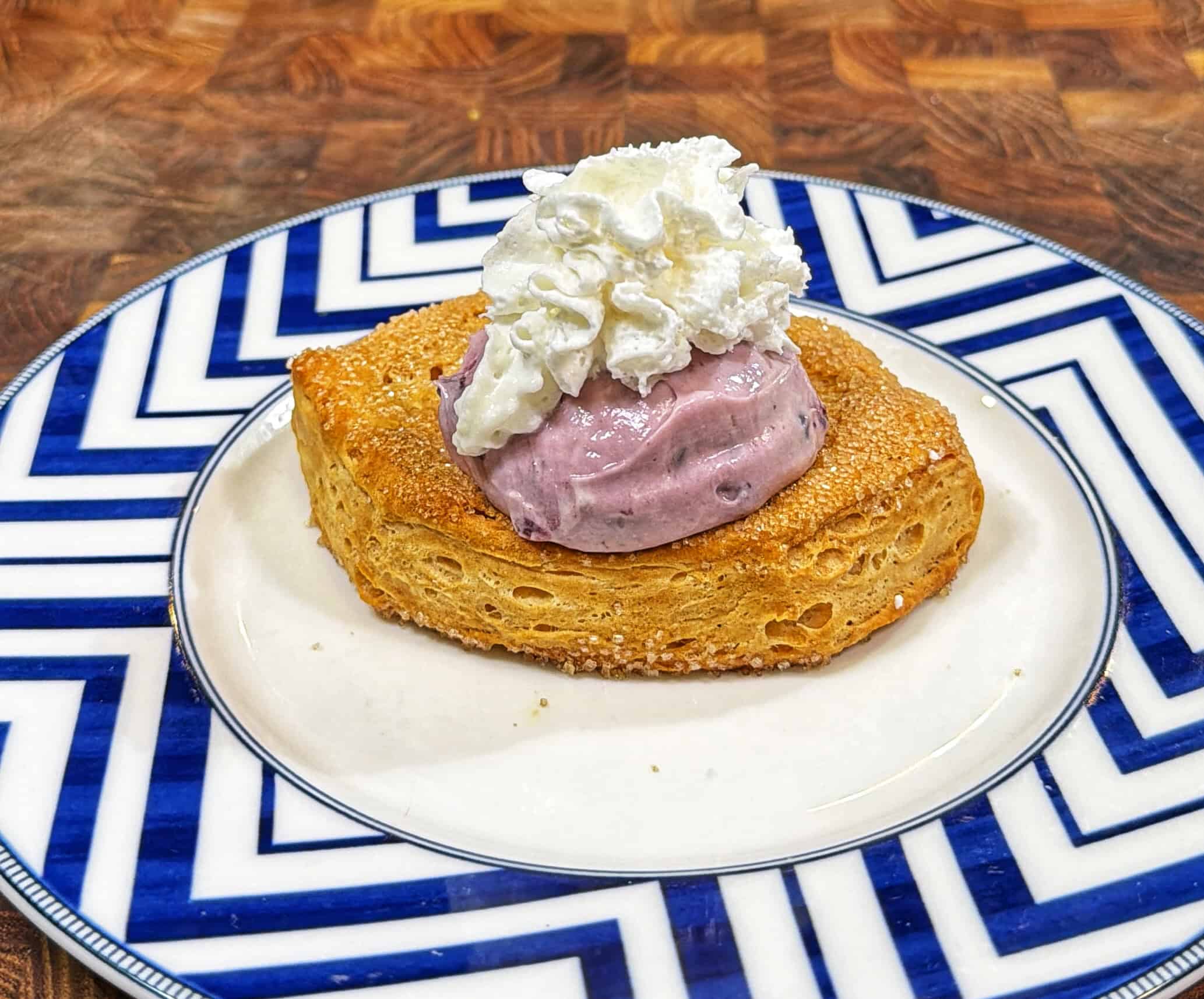 A piece of toasted bread topped with a scoop of purple blueberry cheesecake cream and a swirl of whipped cream, served on a blue and white patterned plate.