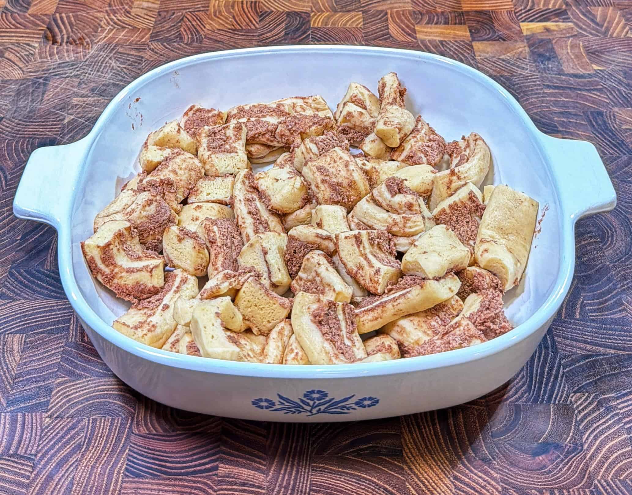 A white casserole dish with blue floral design holds pieces of baked cinnamon roll dough, placed on a wooden surface. The cinnamon rolls appear soft, golden brown, and lightly glazed.