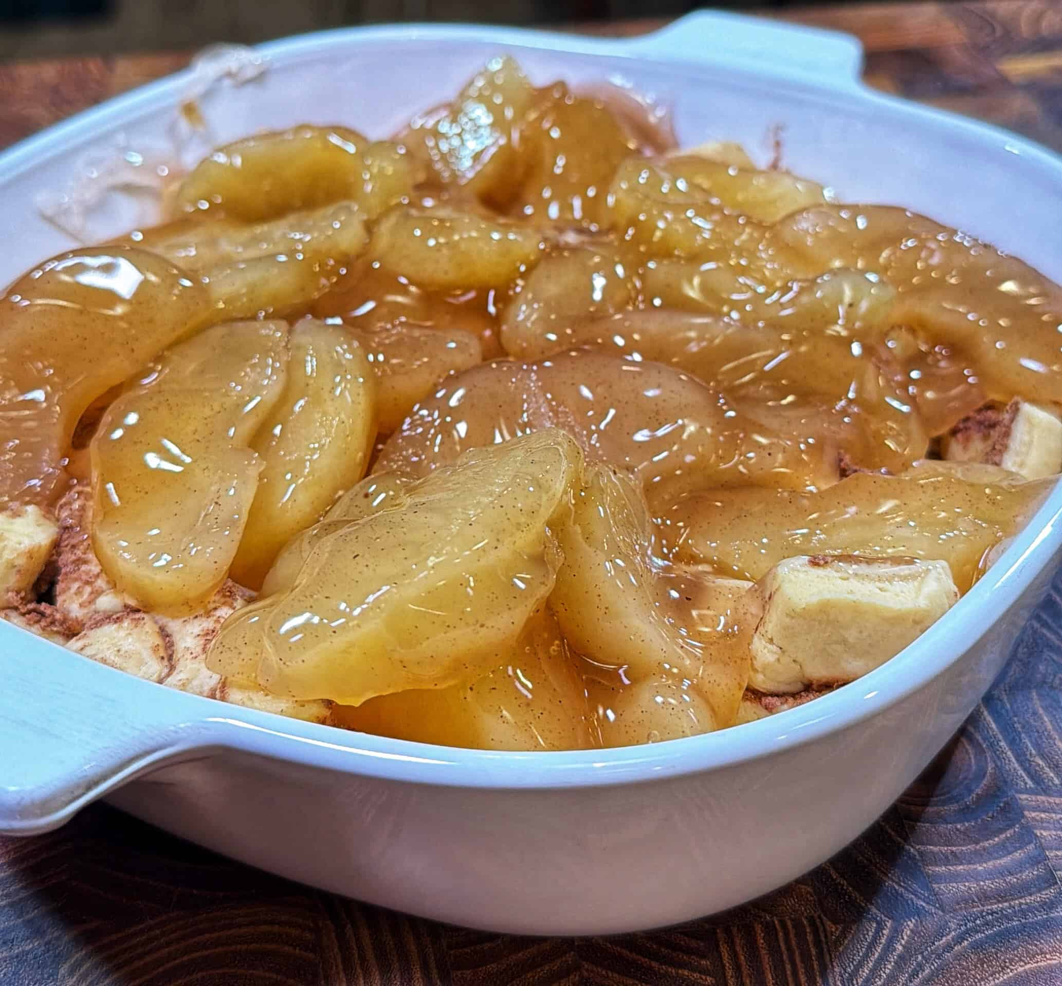 A white baking dish filled with sliced apples coated in a glossy, cinnamon-spiced sauce, ready to be baked, sits on a patterned wooden surface.