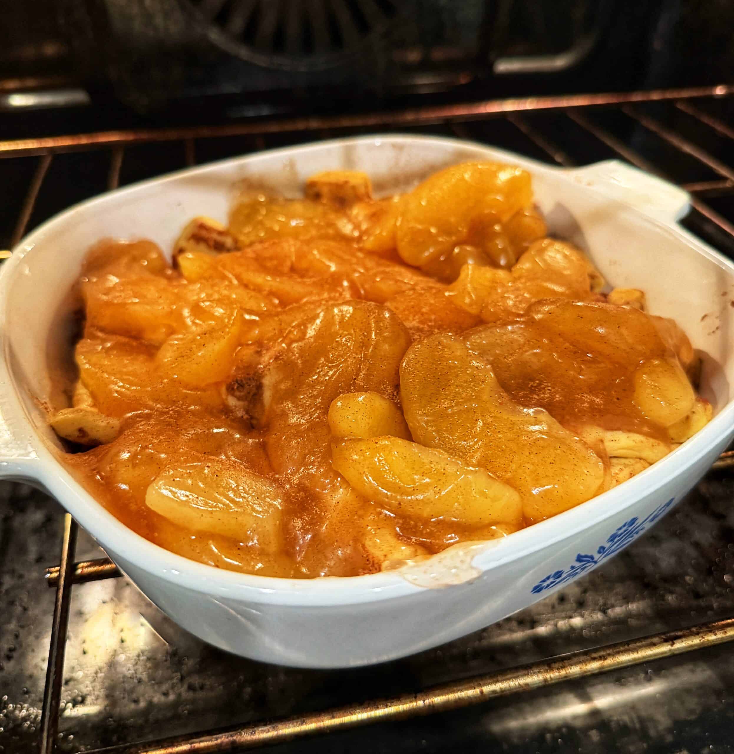 A white ceramic dish filled with bubbling baked apples in a cinnamon sauce sits on an oven rack, with the oven’s heating elements visible in the background.