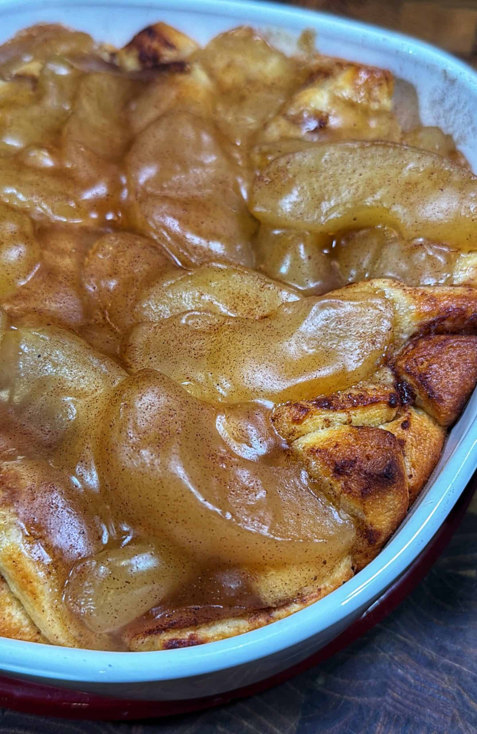 Close-up of a baked dessert in a white and red dish, topped with glossy, cinnamon-spiced apple slices and sauce spread over a golden-brown, doughy base.