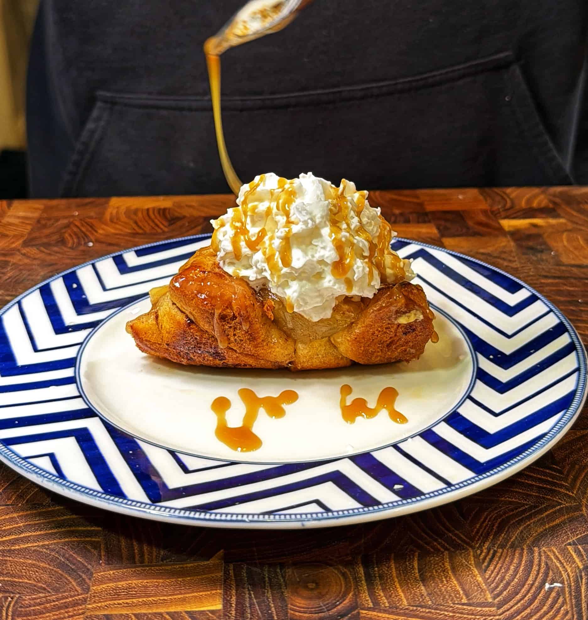 A dessert topped with whipped cream and caramel sauce is served on a blue and white chevron-patterned plate. More caramel sauce is being drizzled from above onto the dessert.