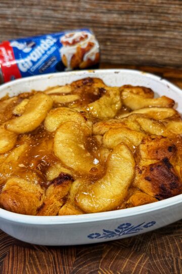 A baked apple cinnamon casserole in a white dish with a blue floral design, with caramelized apple slices on top. In the background, a can of Pillsbury Grands! biscuits is visible.