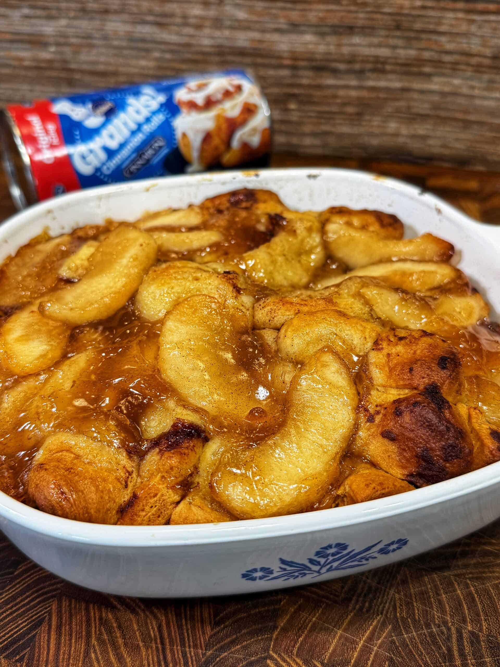 A baked apple cinnamon casserole in a white dish with a blue floral design, with caramelized apple slices on top. In the background, a can of Pillsbury Grands! biscuits is visible.