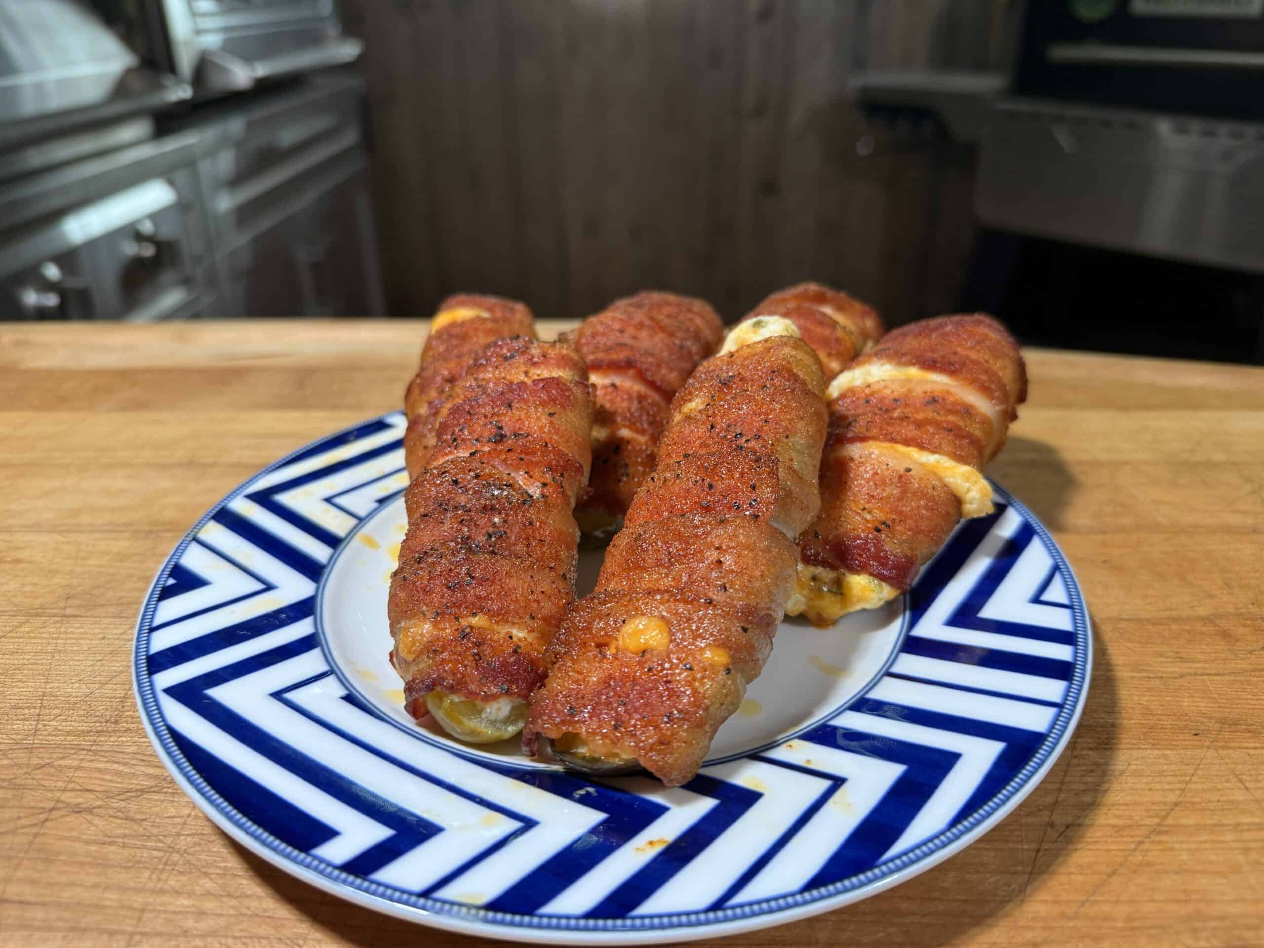 Five bacon-wrapped, cheese-stuffed jalapeño poppers are arranged on a blue and white patterned plate, set on a wooden countertop in a kitchen setting.