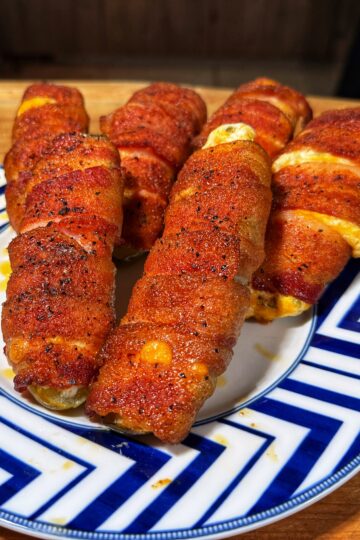 Five crispy, bacon-wrapped, cheese-stuffed jalapeño poppers are arranged on a blue and white patterned plate, sitting on a wooden countertop.