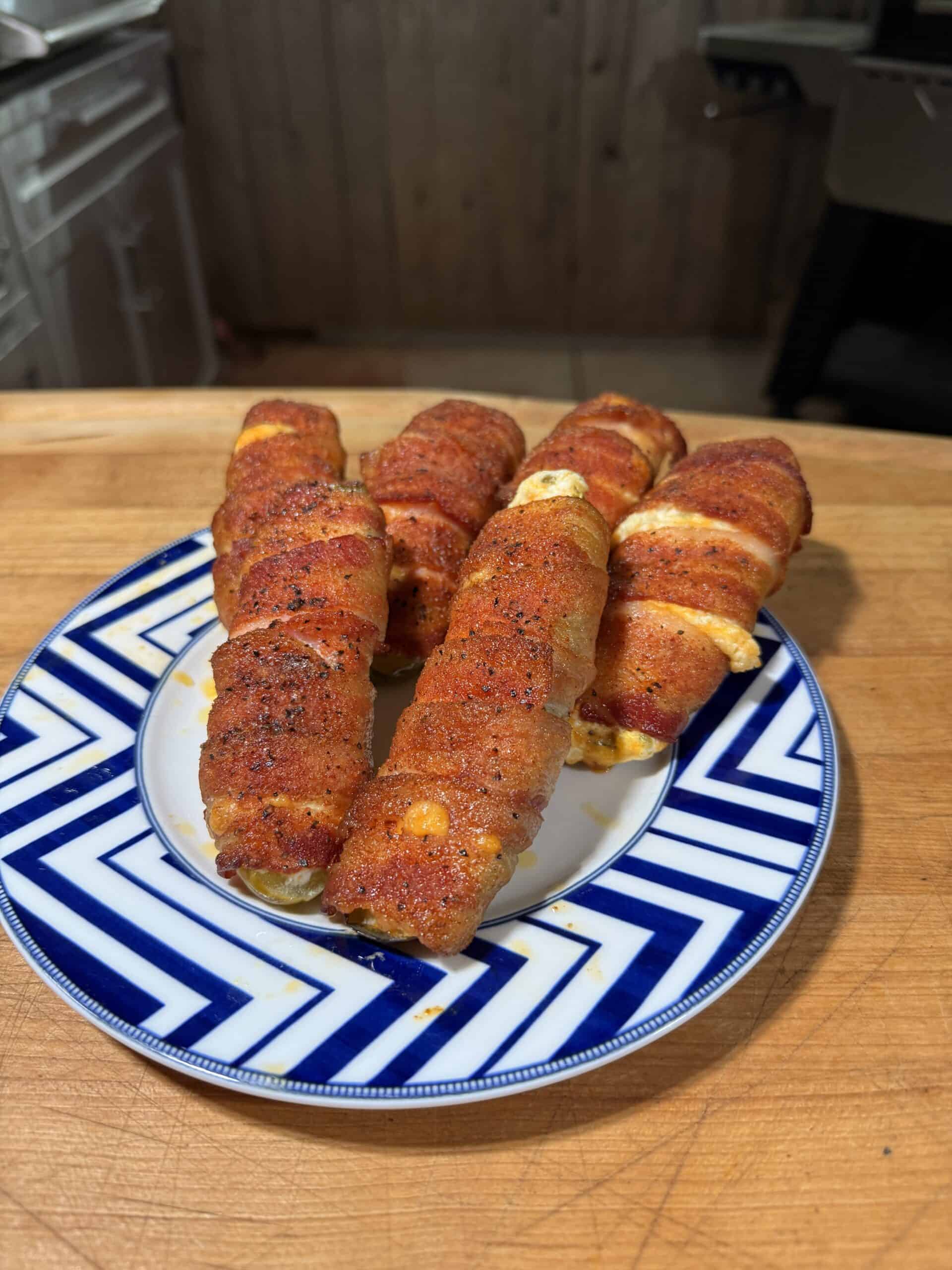 Five bacon-wrapped cheese-stuffed jalapeño poppers, seasoned and baked, are arranged on a blue and white patterned plate on a wooden countertop in a kitchen setting.