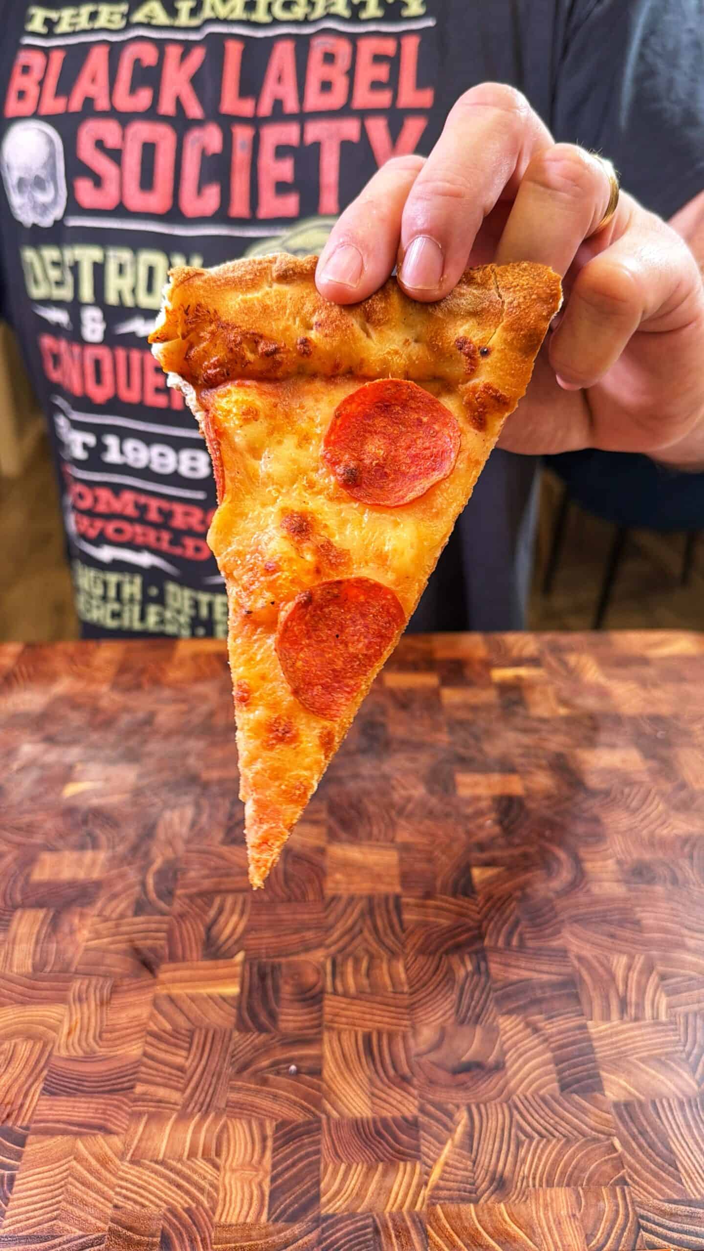 A person wearing a Black Label Society t-shirt holds a slice of pepperoni pizza above a wooden table.