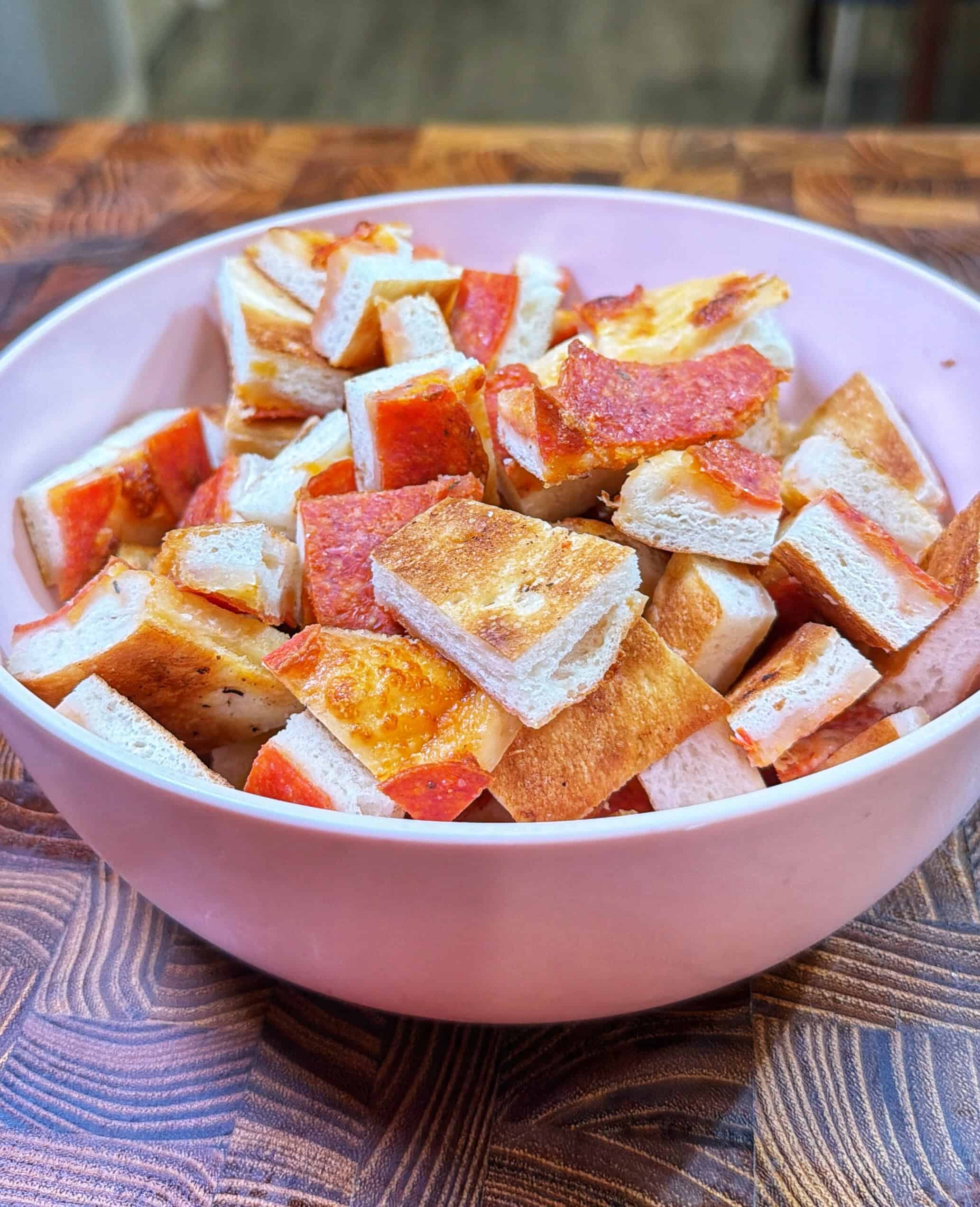 A pink bowl filled with small square pieces of bread topped with pepperoni and melted cheese, set on a wooden surface.