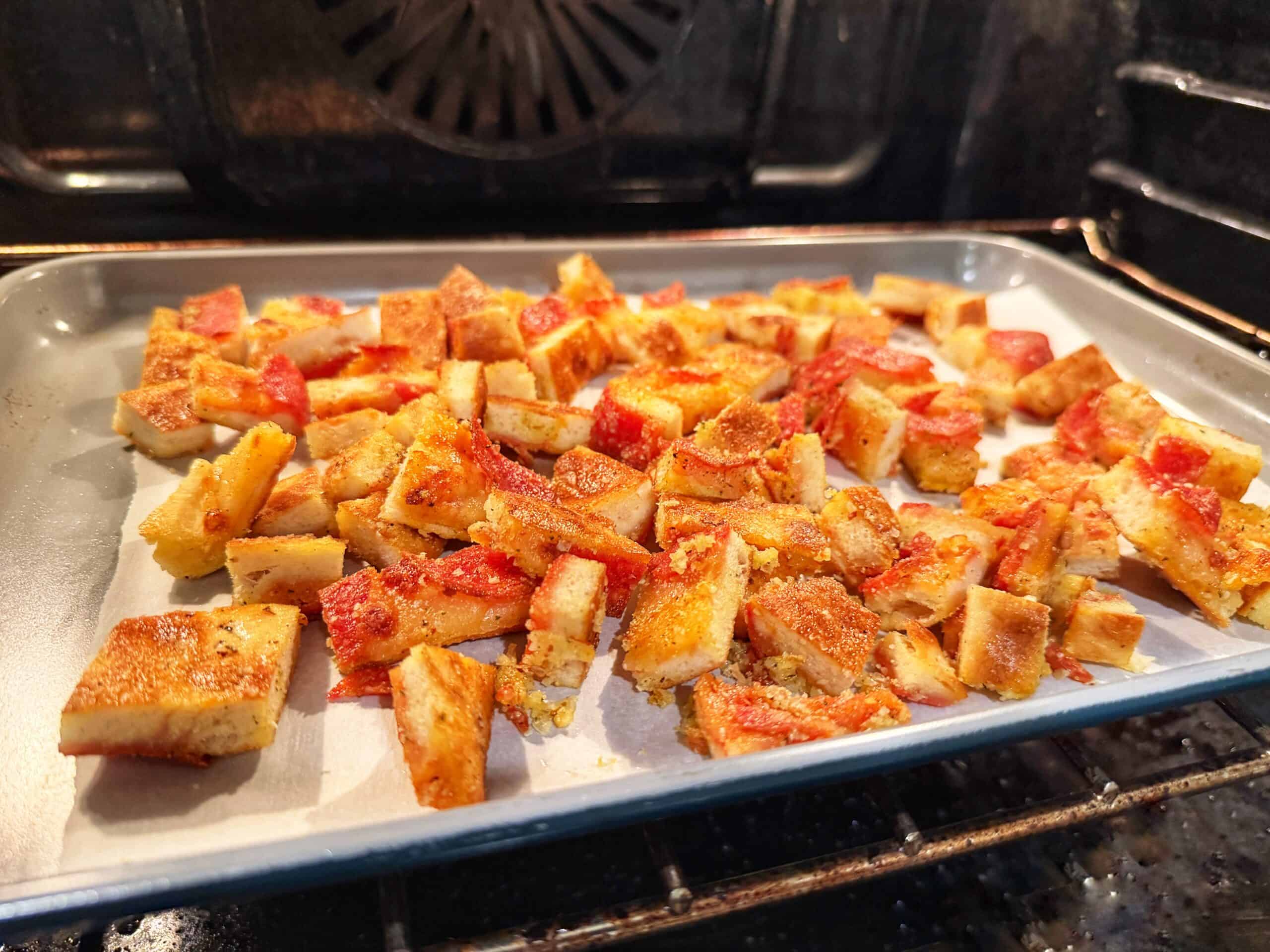 Cubed pieces of pizza croutons with red seasoning are spread on a baking sheet lined with parchment paper, baking inside an oven. The bread cubes appear crispy and toasted.