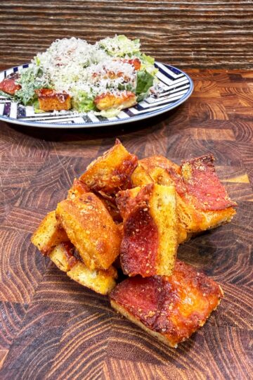 A pile of square pepperoni pizza slices sits on a wooden surface, with a plate of salad and more pizza topped with grated cheese in the background.
