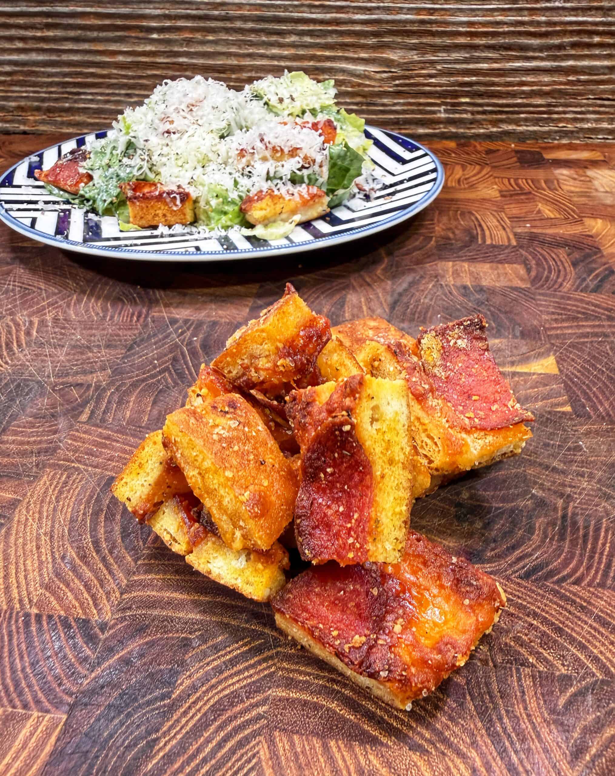 A pile of square pepperoni pizza slices sits on a wooden surface, with a plate of salad and more pizza topped with grated cheese in the background.