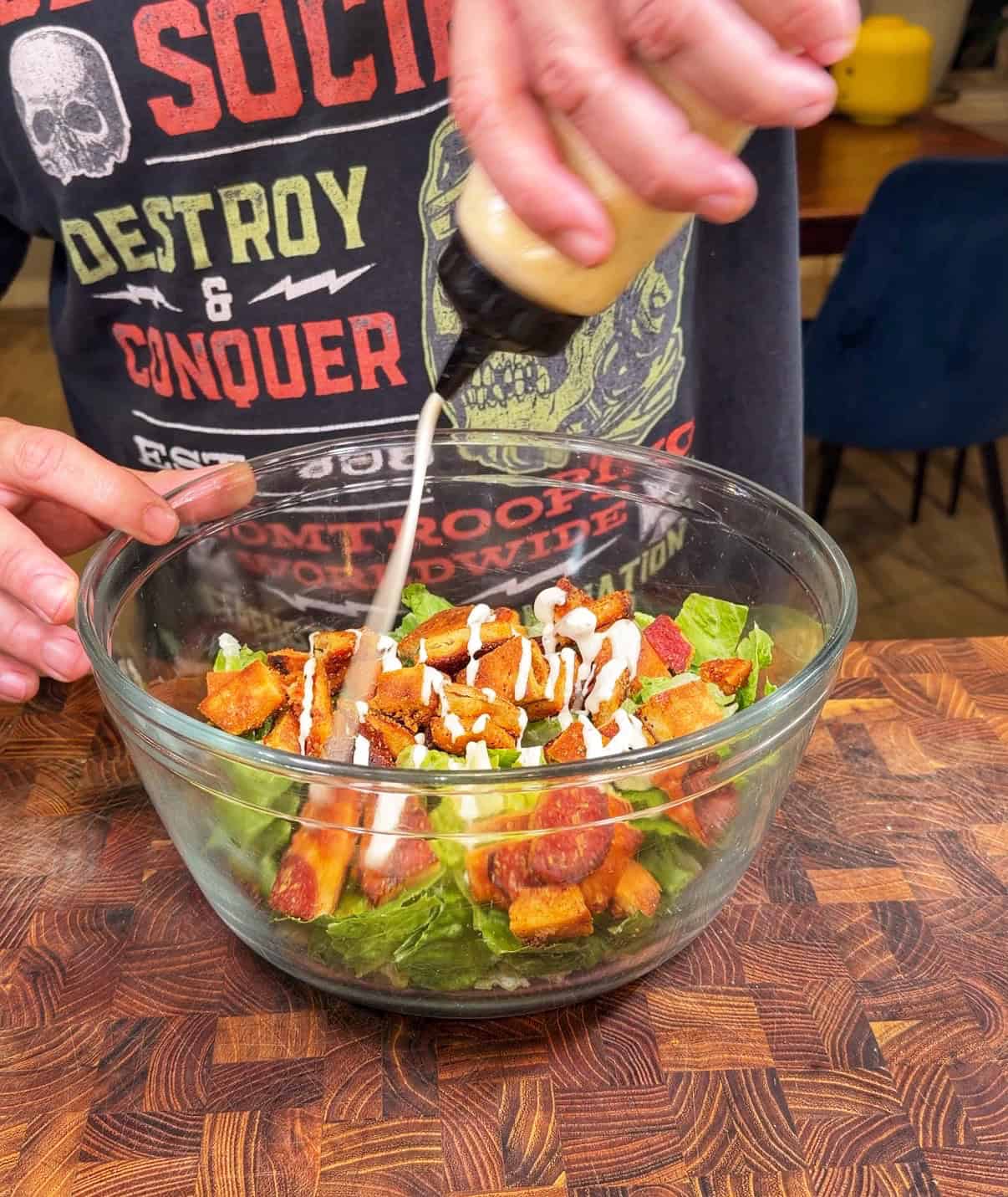 A person pours creamy dressing onto a Caesar salad with croutons and lettuce in a glass bowl, standing at a wooden countertop. They are wearing a graphic t-shirt with text and a skull design.