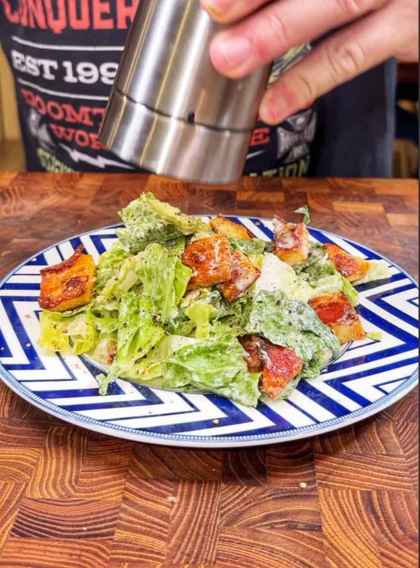A person grinds pepper onto a plate of Caesar salad with croutons and lettuce on a blue and white patterned plate, set on a wooden table.