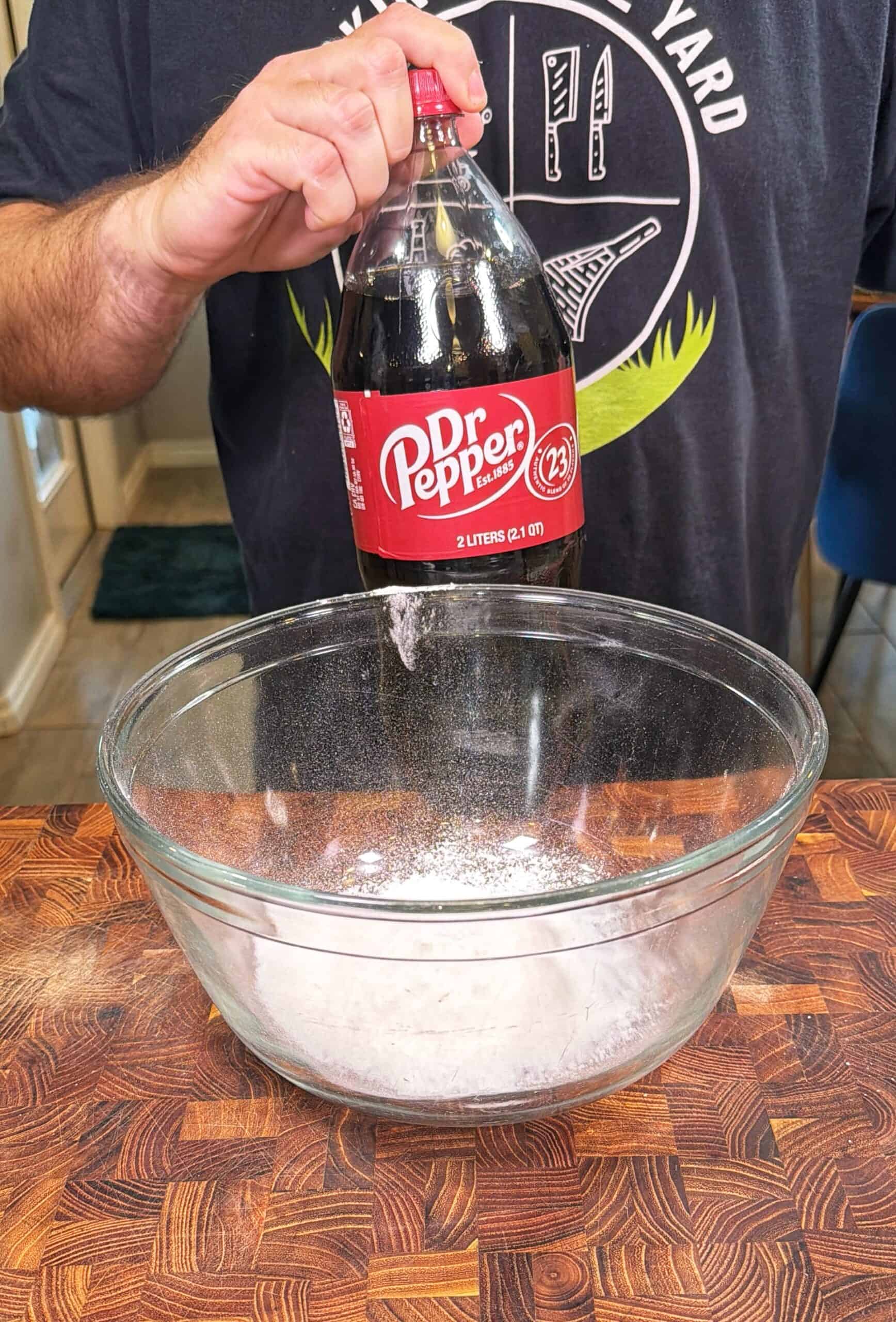 A person holds a 2-liter bottle of Dr Pepper over a large glass bowl filled with white powder, preparing to pour it in. The scene is set on a wooden counter in a kitchen.