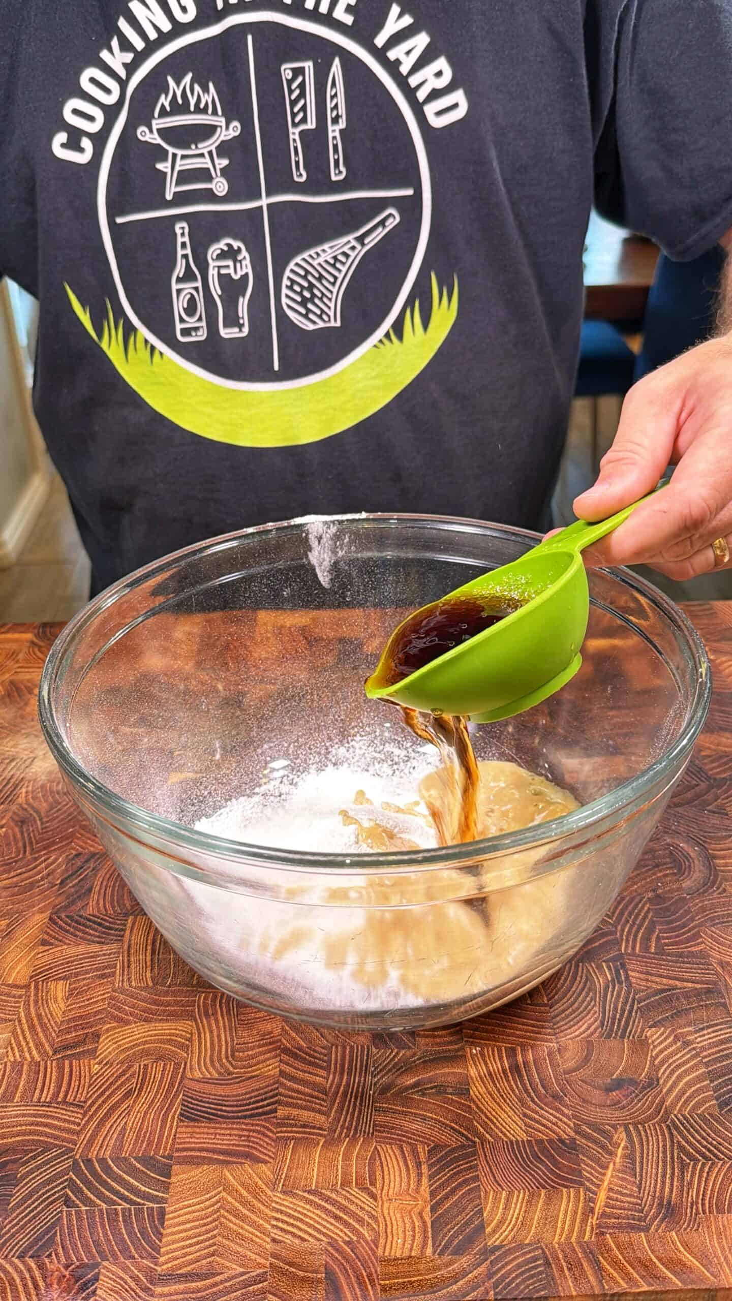 A person in a Cooking in the Yard shirt pours a dark liquid from a green measuring cup into a glass mixing bowl containing a beige and white mixture on a wooden countertop.