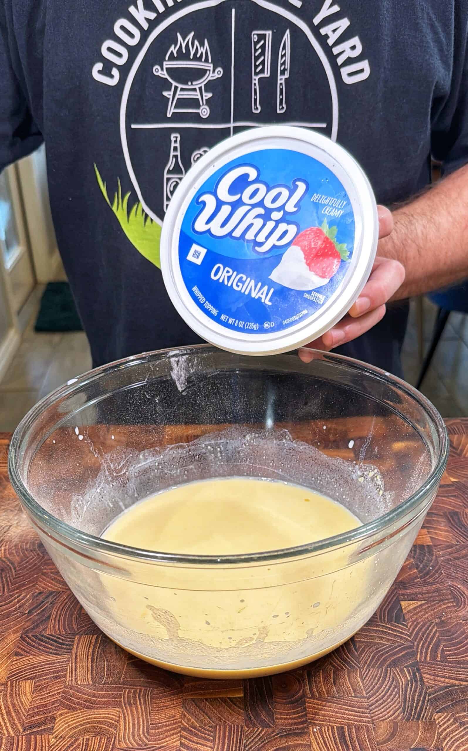 A person holds a container of Cool Whip Original above a glass bowl filled with a creamy yellow mixture, on a wooden countertop. The person is wearing a shirt with cooking-themed graphics.