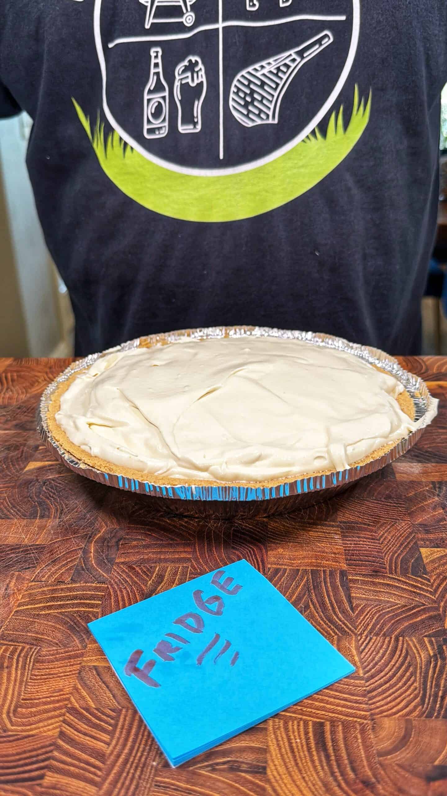 A pie with a creamy white topping sits on a wooden surface. A blue sticky note labeled FUDGE is in front of the pie. A person in a dark T-shirt with white graphics stands behind the pie.