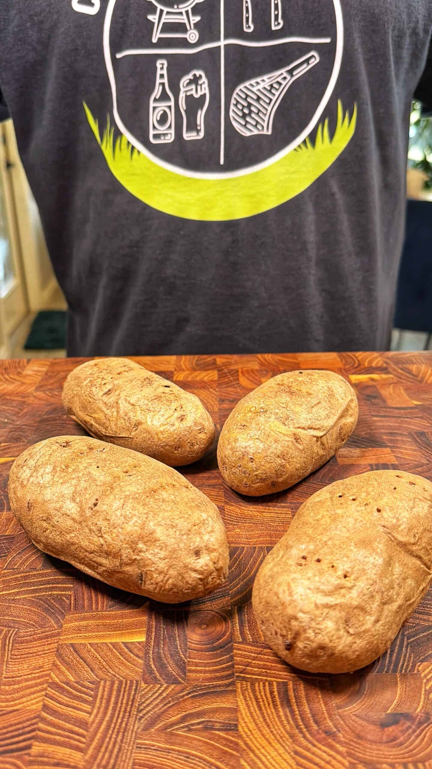 Four baked potatoes rest on a wooden cutting board. In the background, a person wearing a dark t-shirt with a white and green graphic stands behind the board.
