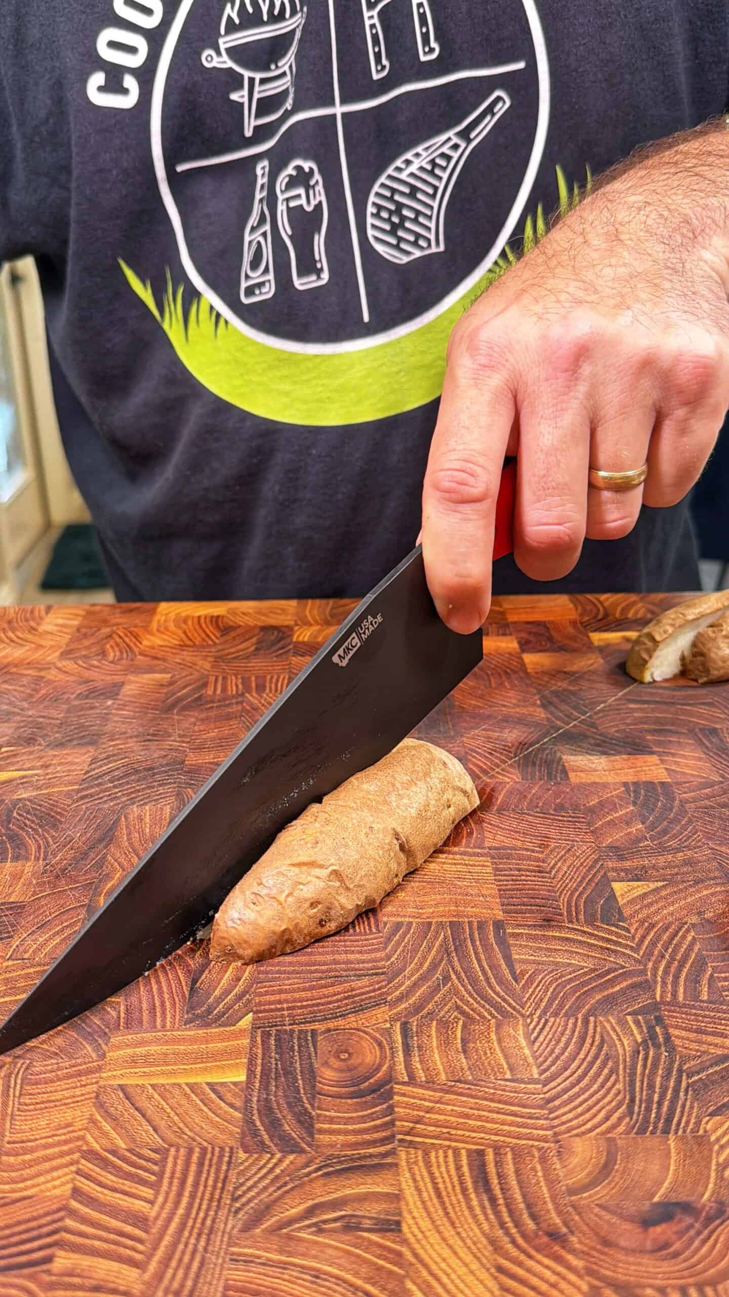 A person wearing a dark shirt is slicing a small potato with a large black knife on a patterned wooden cutting board. Only their hand and part of the torso are visible.