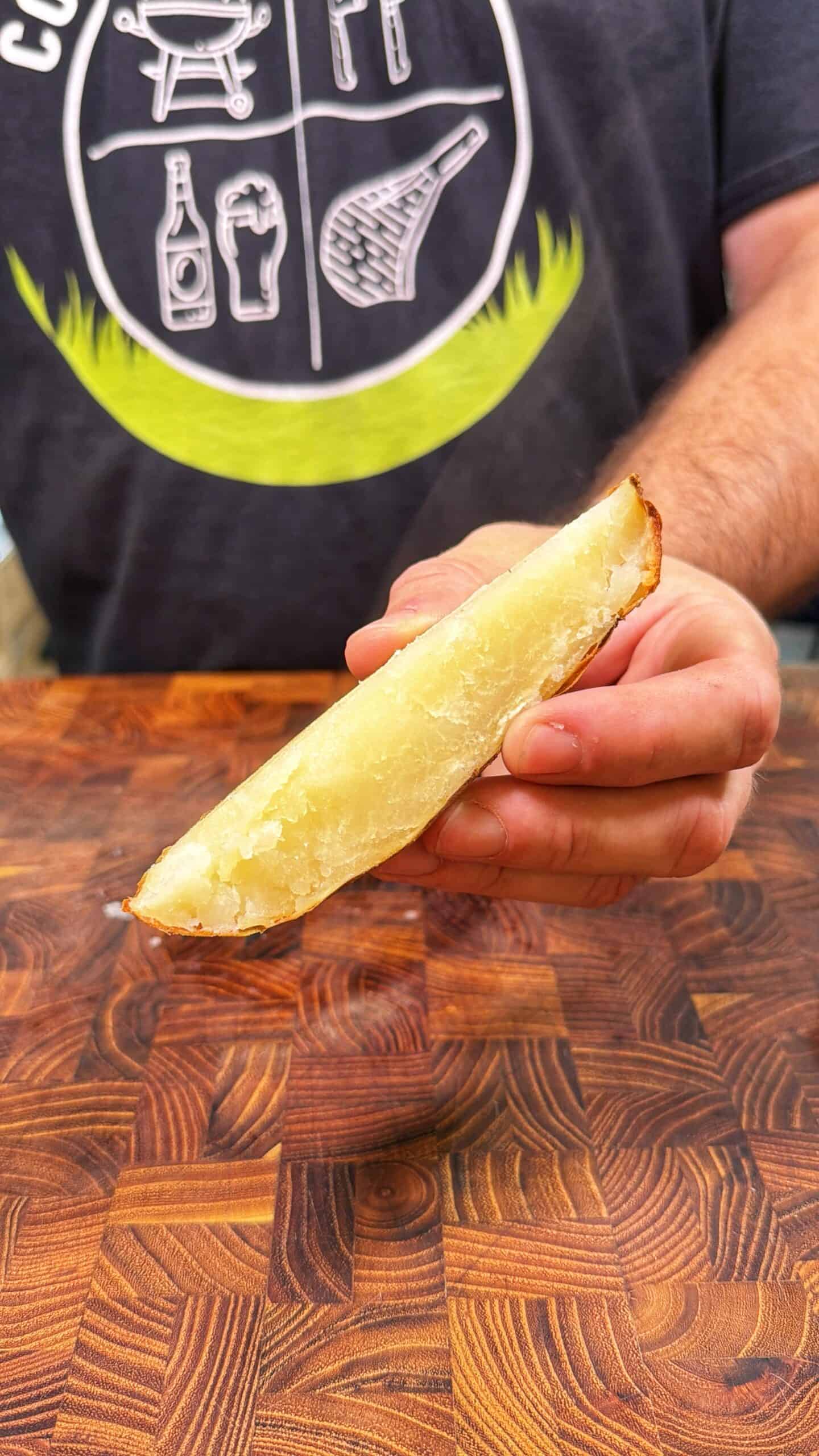 A person holds a large, plain potato wedge over a wooden surface. The person is wearing a dark shirt with white food-related graphics and green accents.