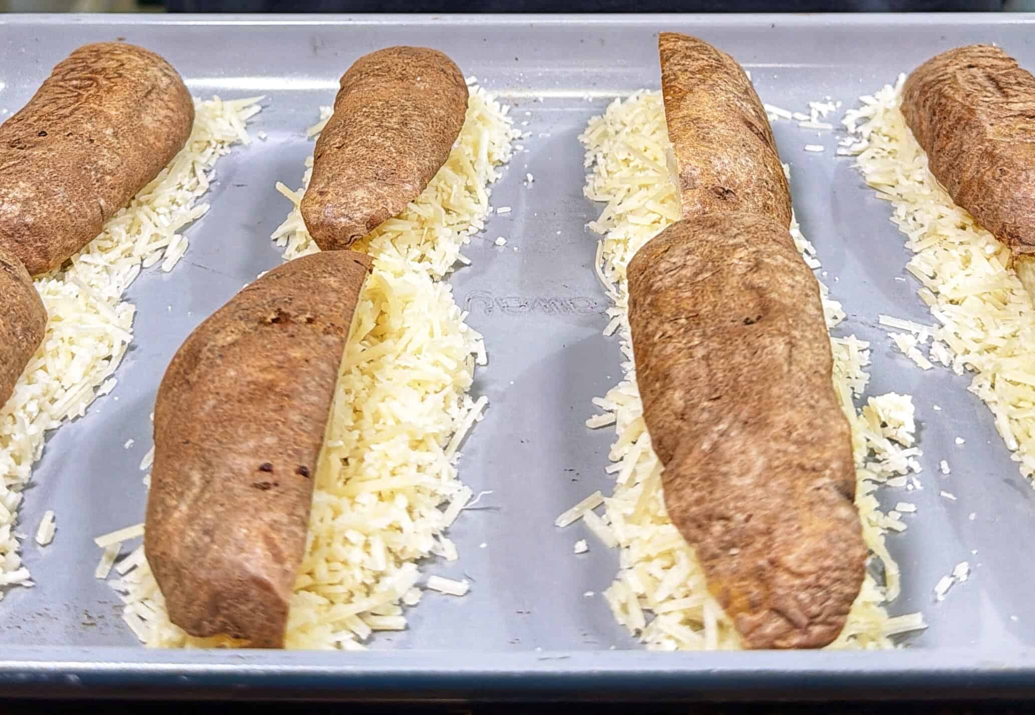 Baked potato halves placed cut-side up on a baking tray, each resting on a bed of shredded cheese, ready for further cooking or topping.