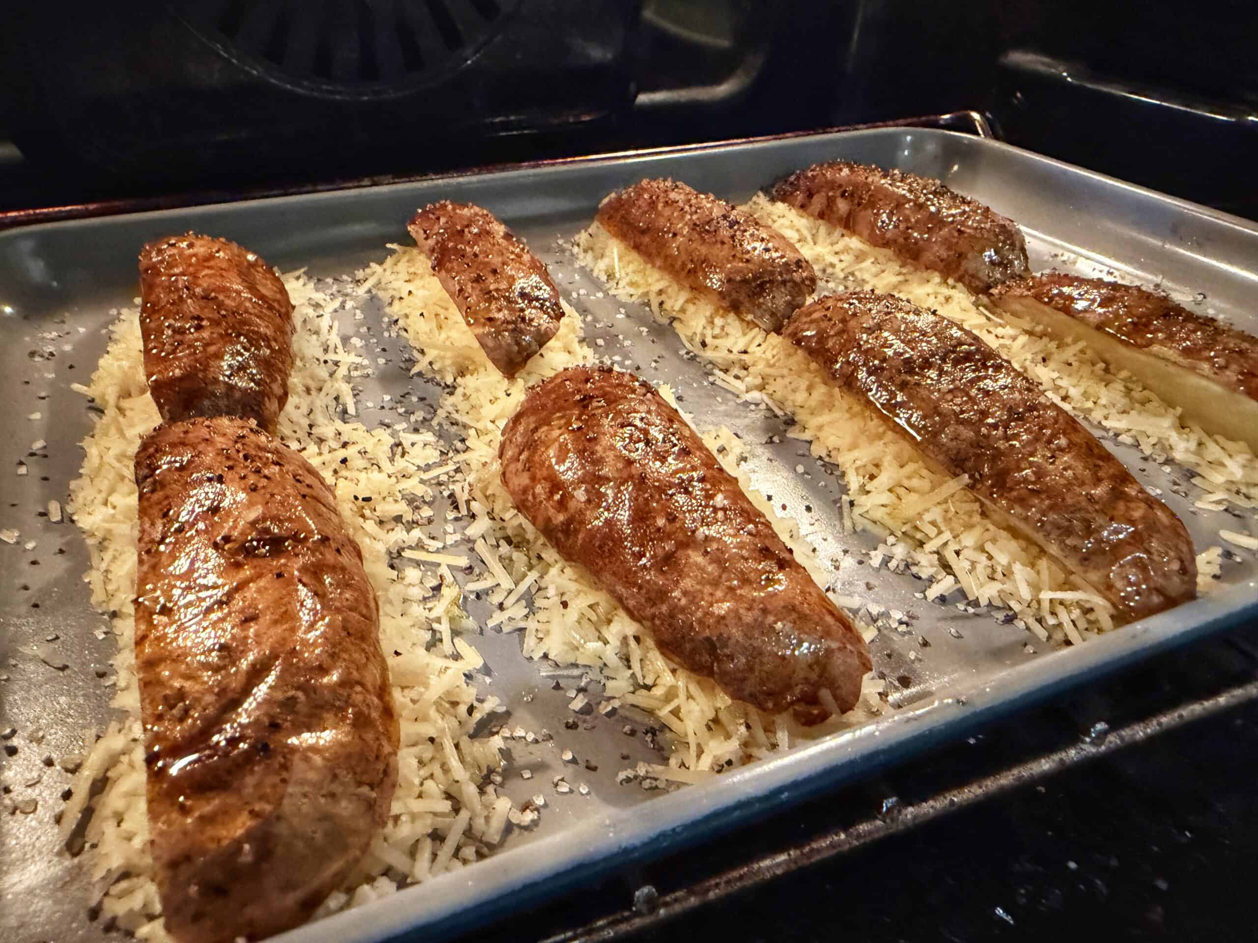 Several potato wedges, seasoned and placed cut-side down on shredded cheese, are baking on a metal tray in an oven. The cheese is beginning to melt and brown around the potatoes.
