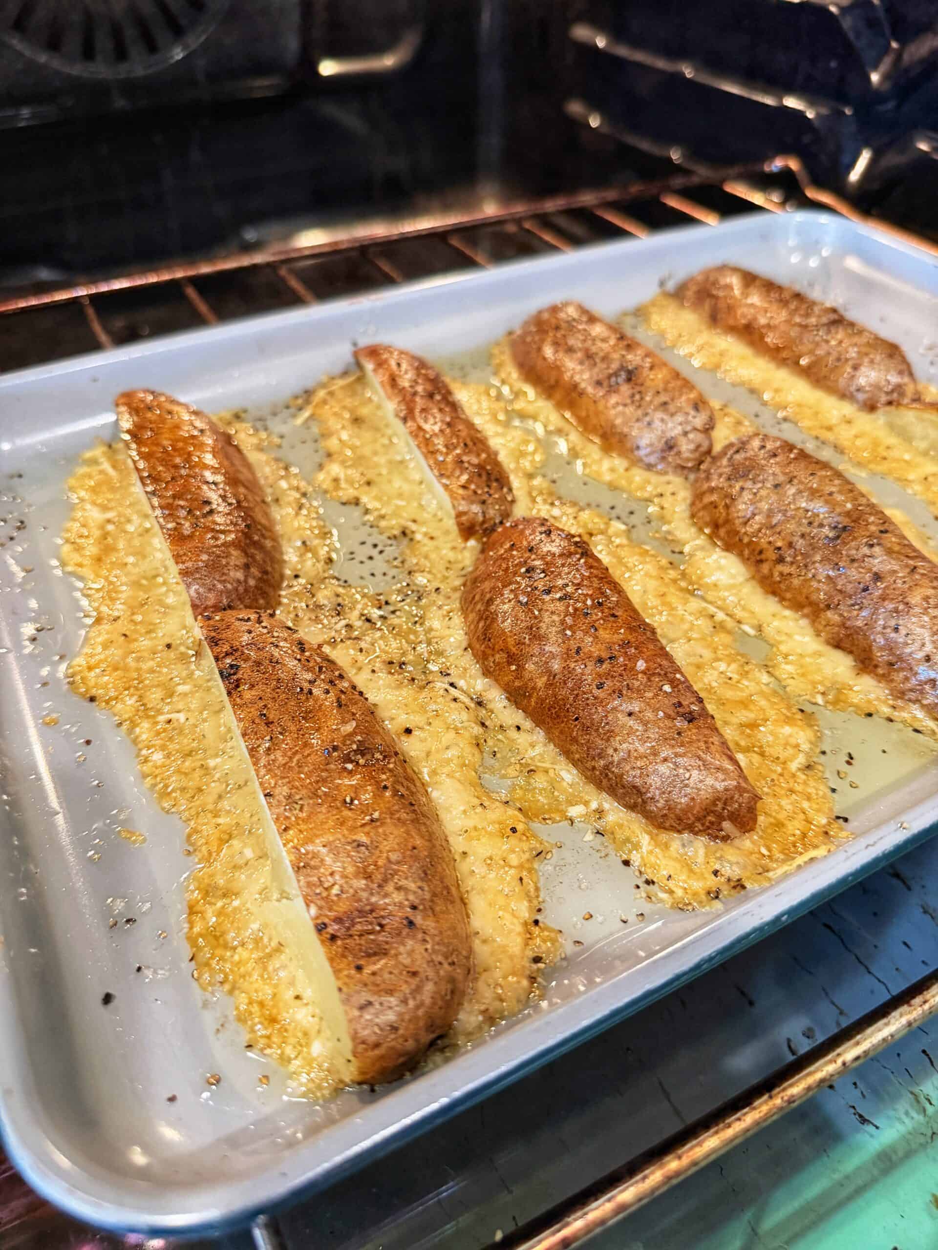 Potato wedges coated in seasoning and cheese are baking on a tray in the oven. Melted cheese is bubbling and spreading around the potatoes, giving a crispy, golden appearance.