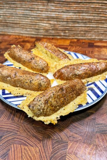 Five sausages are placed in taco-shaped crispy cheese shells on a blue and white striped plate, sitting on a patterned wooden surface with a textured wooden background.