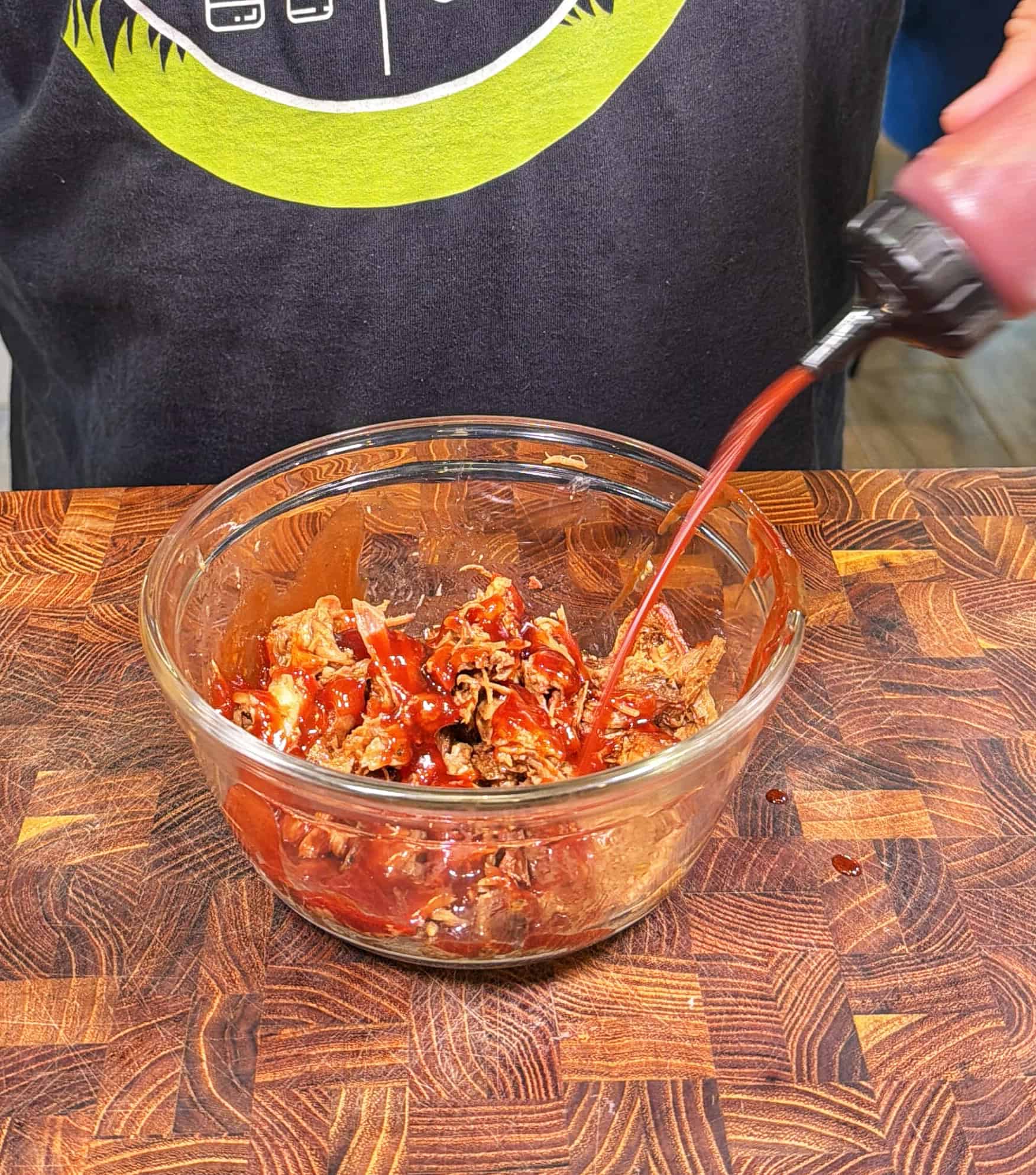 A person pours barbecue sauce from a squeeze bottle onto shredded meat in a glass bowl, which is sitting on a wooden cutting board.