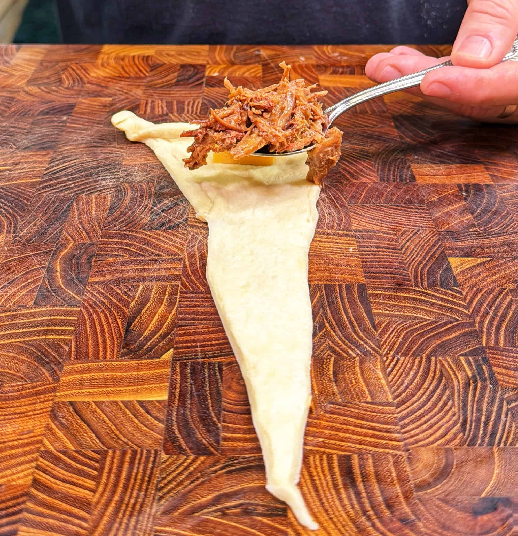 A hand holds a spoon placing shredded meat onto a triangular piece of raw dough on a wooden cutting board.
