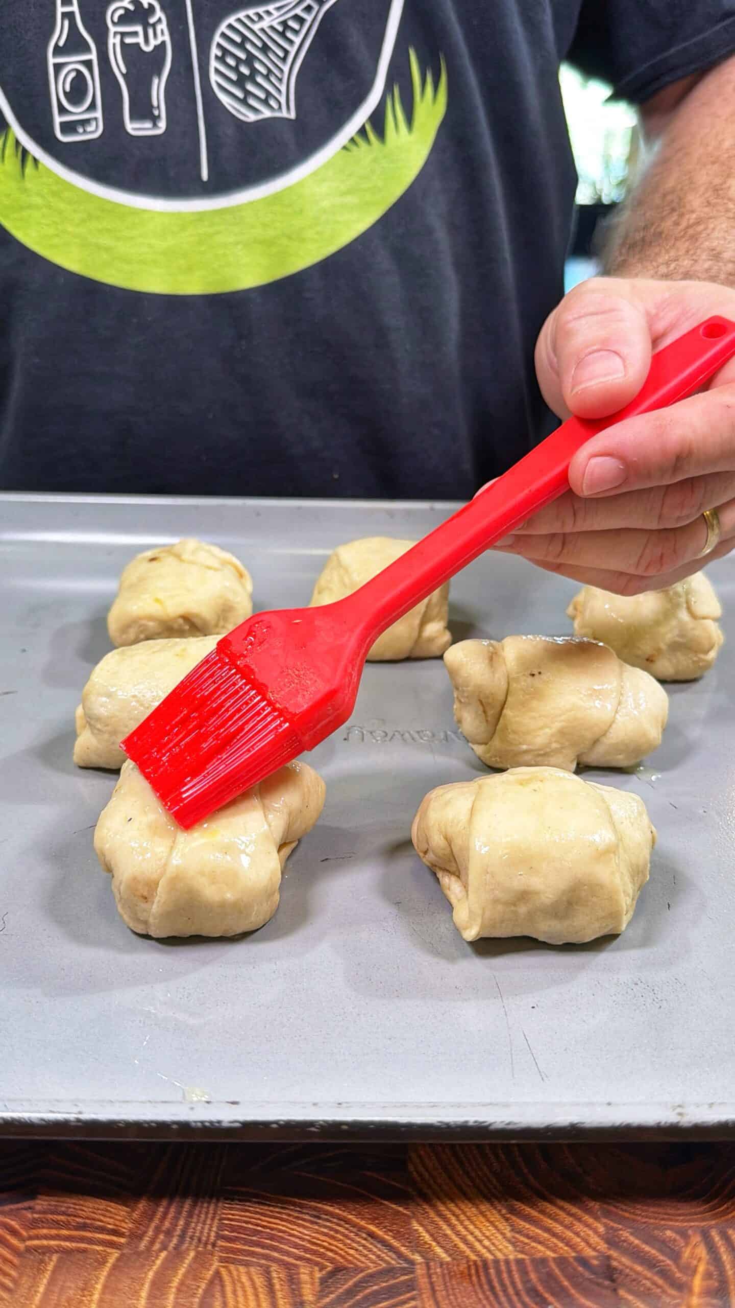 A hand uses a red pastry brush to apply a glaze to uncooked dough balls on a baking sheet. The person is wearing a dark shirt with white graphic designs near the top of the image.