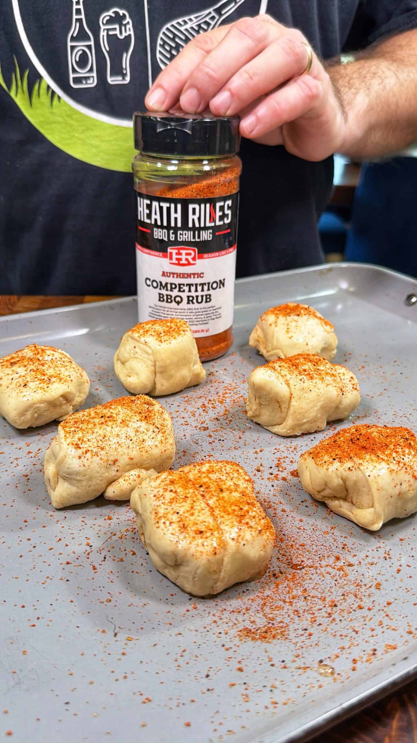 A hand sprinkles Heath Riles Competition BBQ Rub onto seven pieces of uncooked, wrapped dough on a baking tray. The person wears a dark shirt with barbecue-themed graphics in the background.