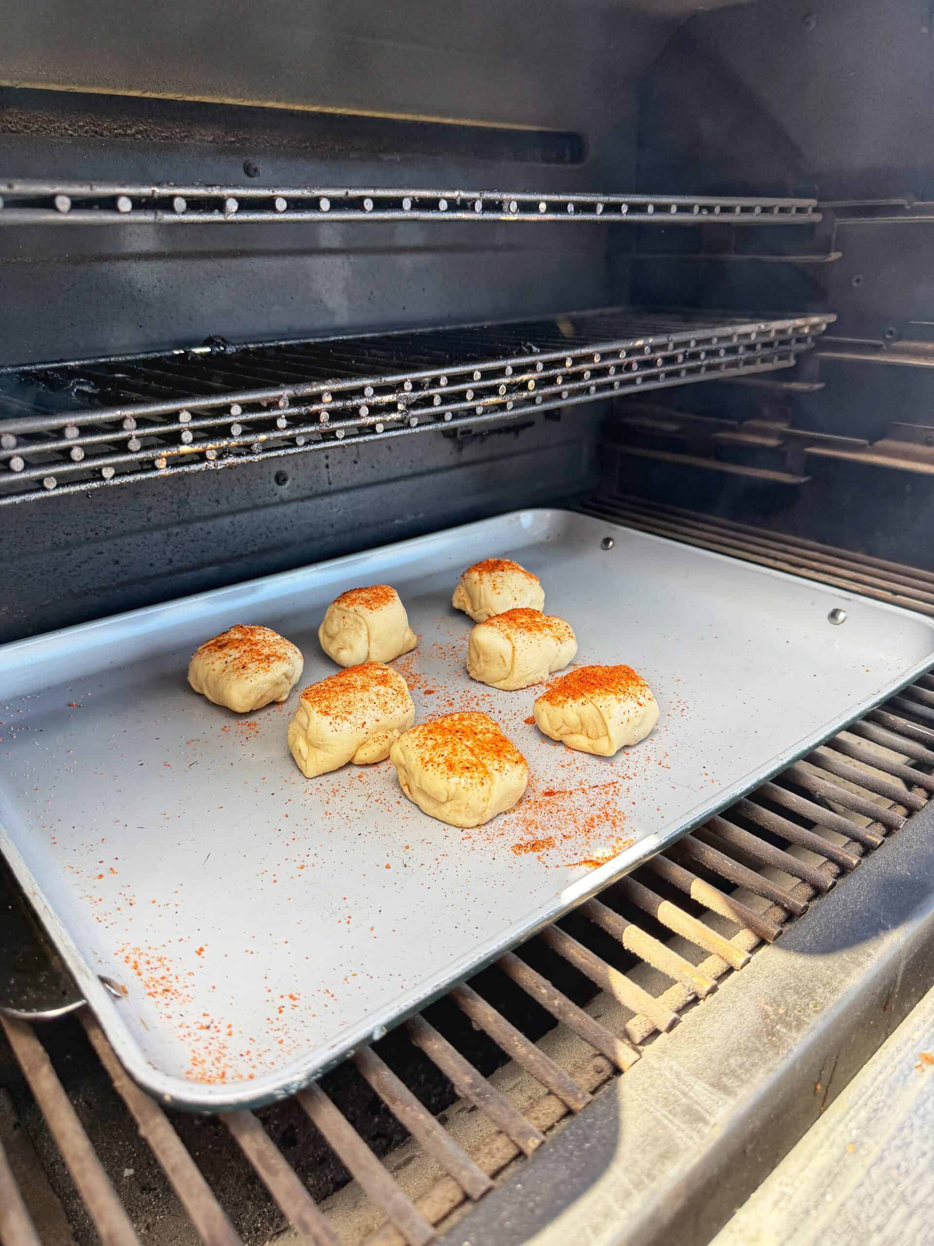 A baking tray with seven dough balls sprinkled with seasoning sits on a rack inside an oven, ready to be baked. The oven interior and additional empty racks are visible in the background.