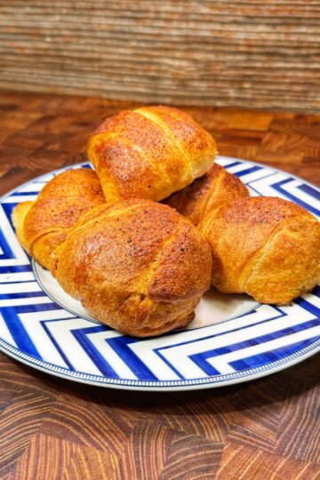 A blue and white patterned plate holds four golden-brown, freshly baked bread rolls on a wooden surface with a textured brown background.