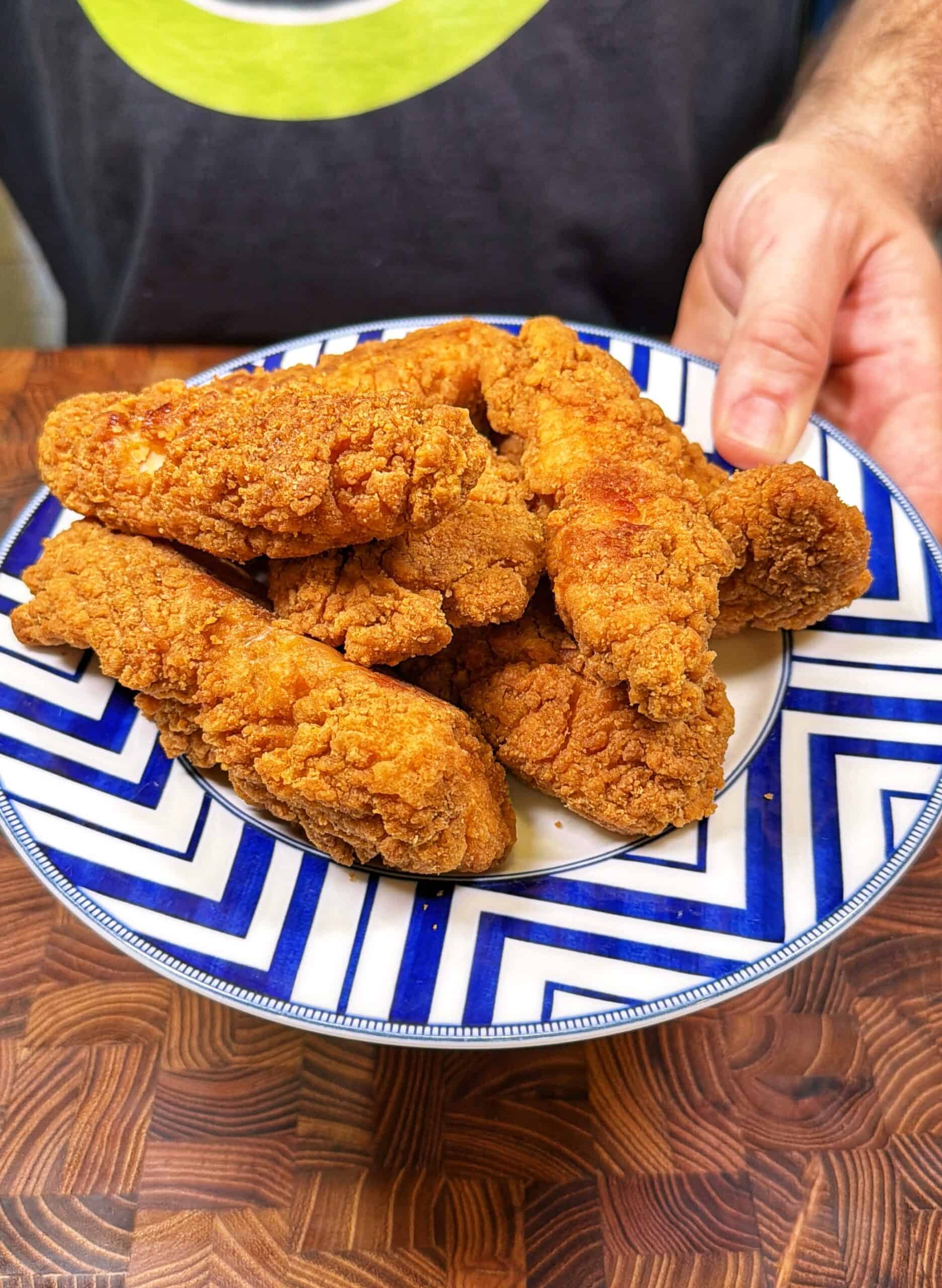 A person holds a blue and white plate filled with several pieces of crispy, golden-brown fried chicken tenders over a wooden surface.