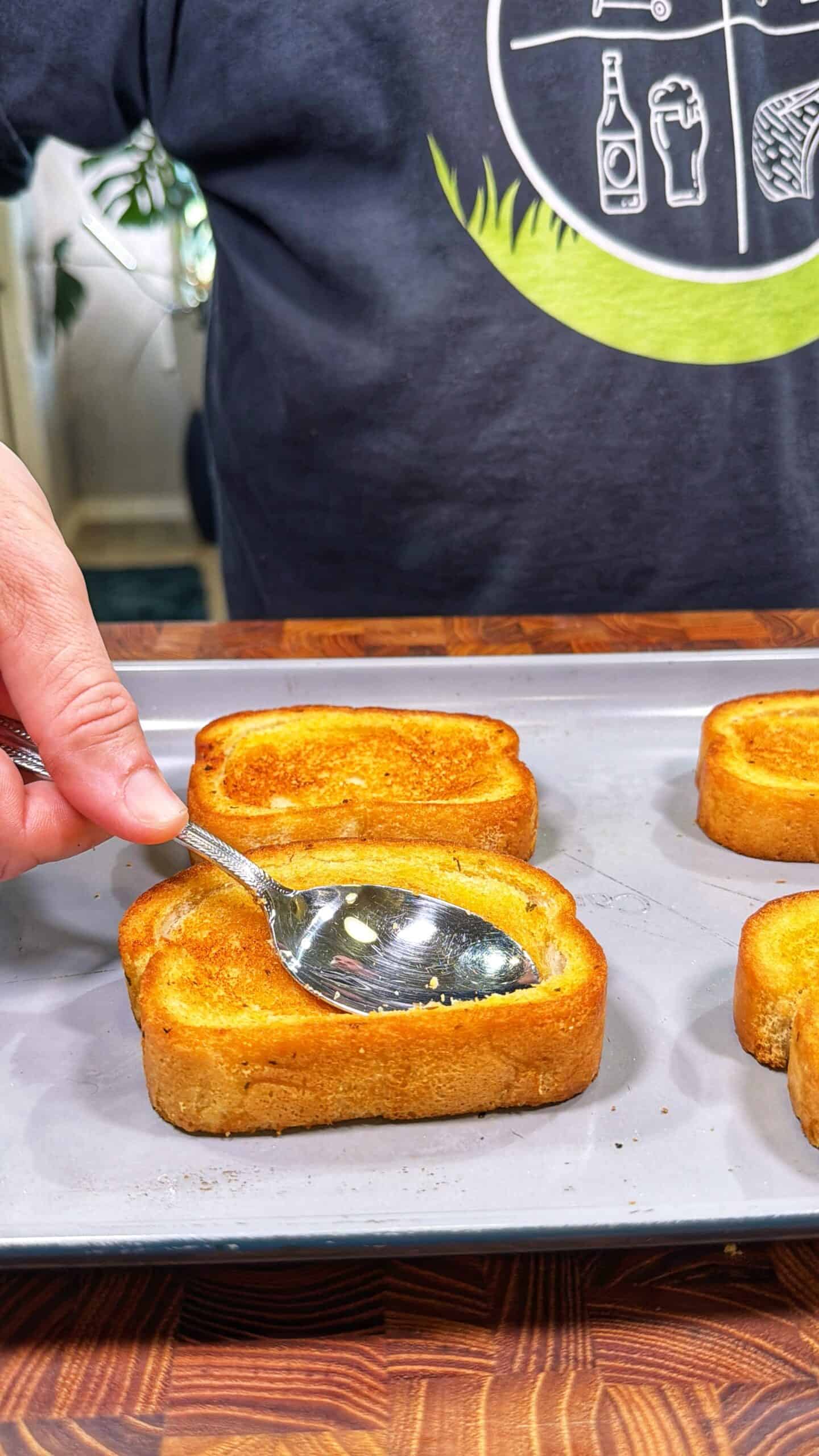 A person uses a spoon to press into the center of a slice of toasted bread on a baking sheet, while other pieces of toast are nearby. The person is wearing a dark shirt with white graphics.