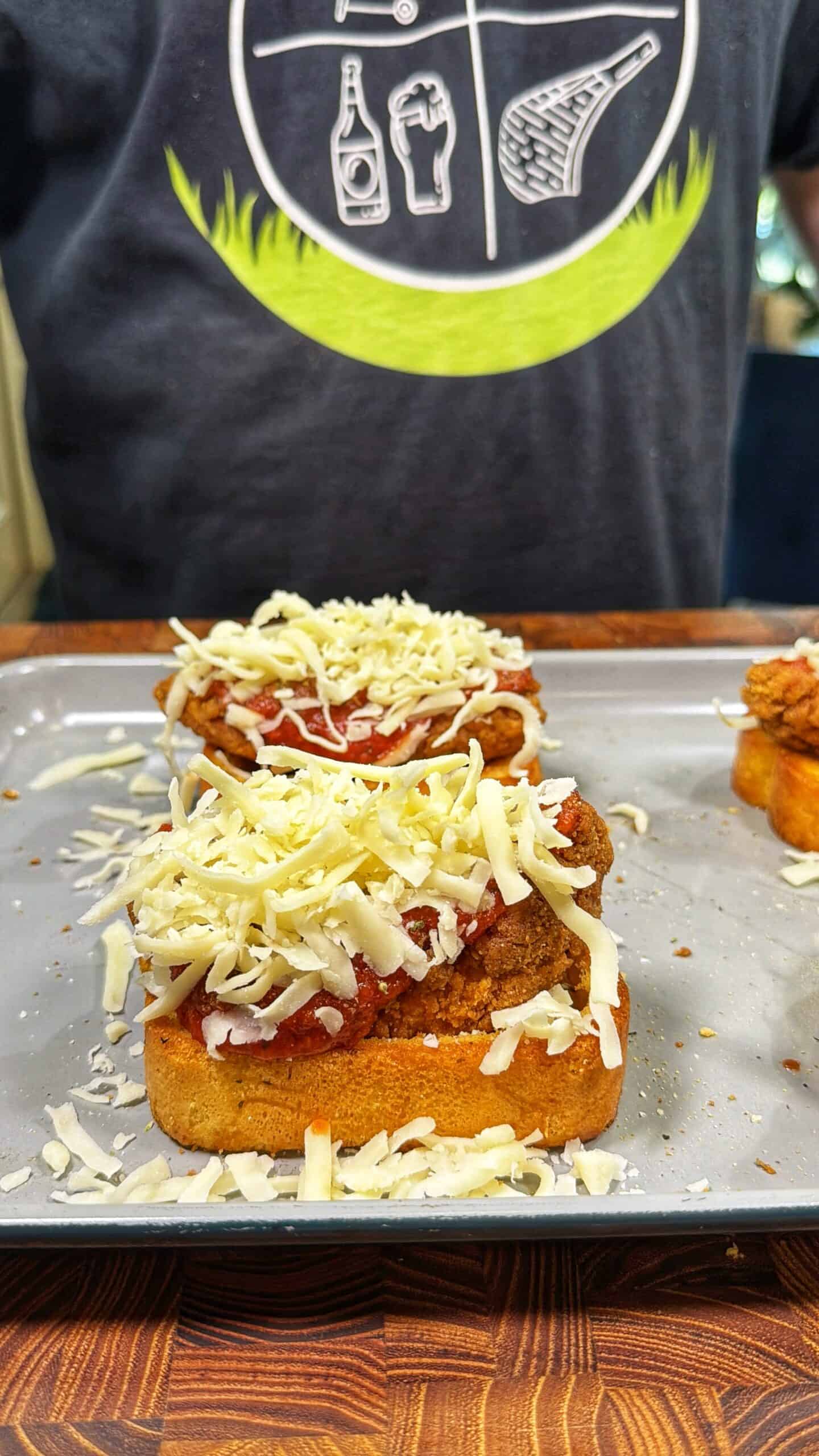 Two slices of bread topped with tomato sauce, breaded fried chicken, and shredded cheese sit on a baking tray. A person wearing a dark shirt with food illustrations stands behind the tray.