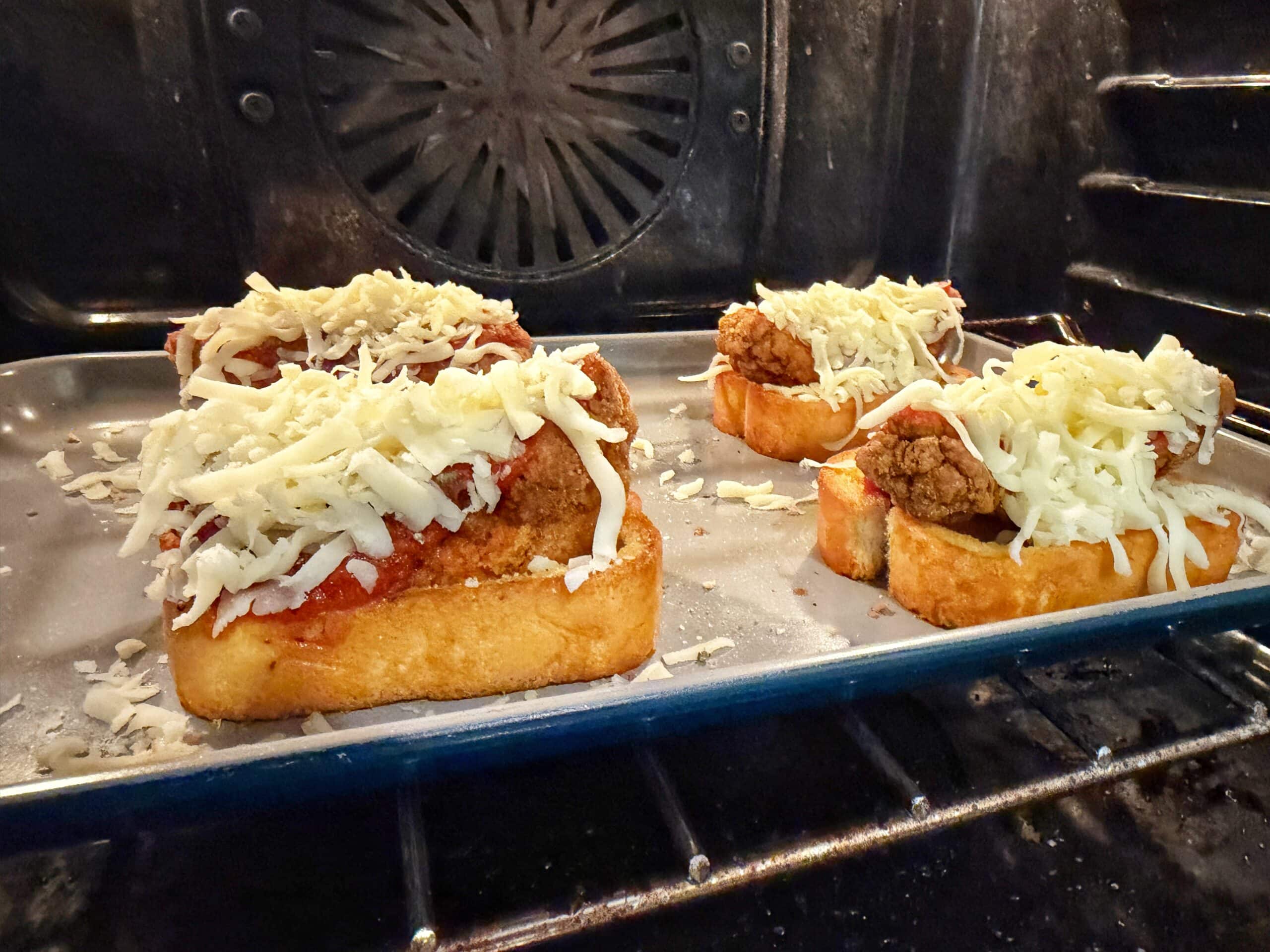 Three slices of bread topped with meatballs, tomato sauce, and shredded cheese are baking on a tray in an oven.