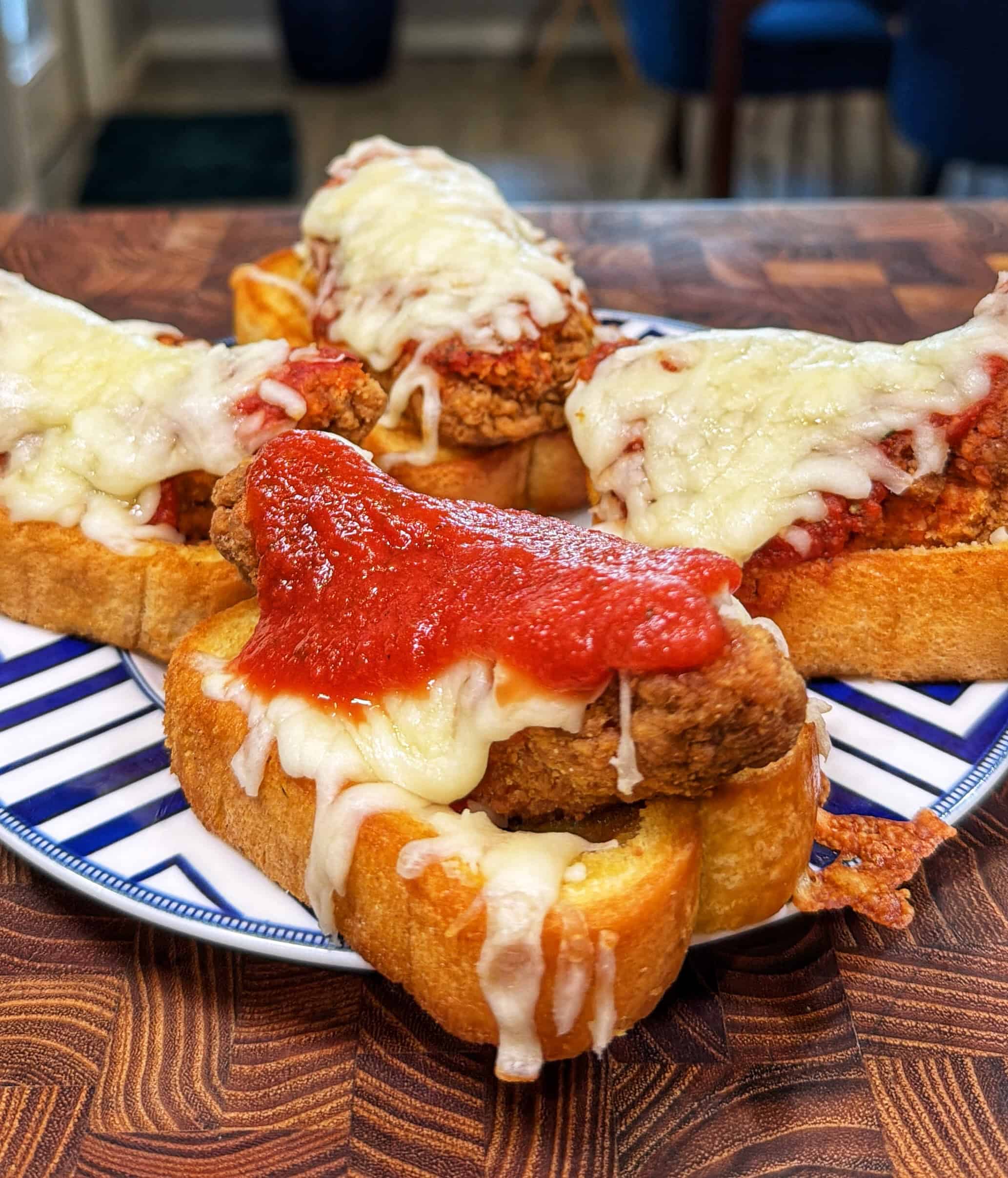Four slices of toasted bread topped with crispy fried chicken, marinara sauce, and melted mozzarella cheese are arranged on a blue and white plate on a wooden table.