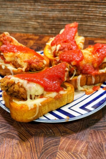 Four slices of toasted bread topped with fried chicken, melted cheese, and marinara sauce are arranged on a patterned plate on a wooden surface.