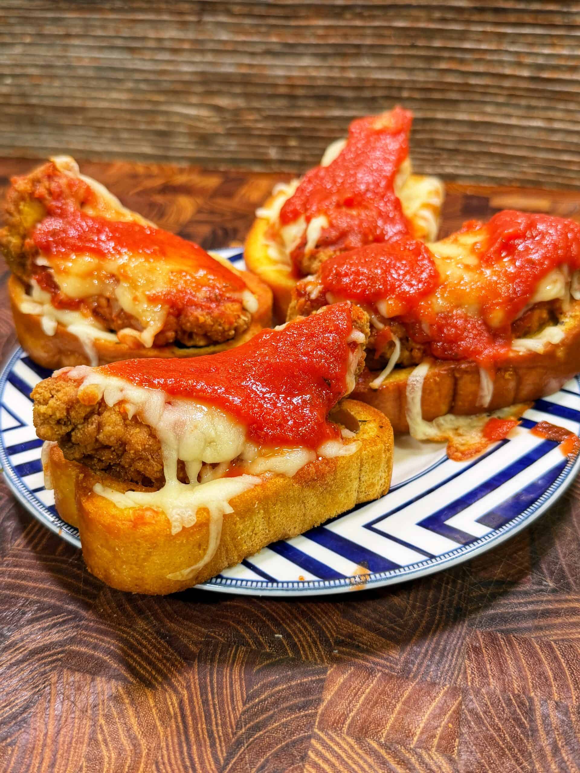 Four slices of toasted bread topped with fried chicken, melted cheese, and marinara sauce are arranged on a patterned plate on a wooden surface.