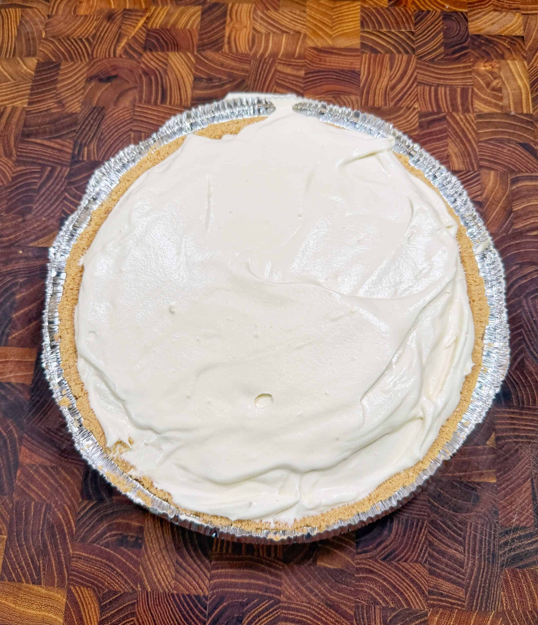 A Dr Pepper Pie with a smooth, creamy white filling sits in a foil pie tin on a wooden, patterned cutting board. The pie has a graham cracker crust and appears to be homemade.