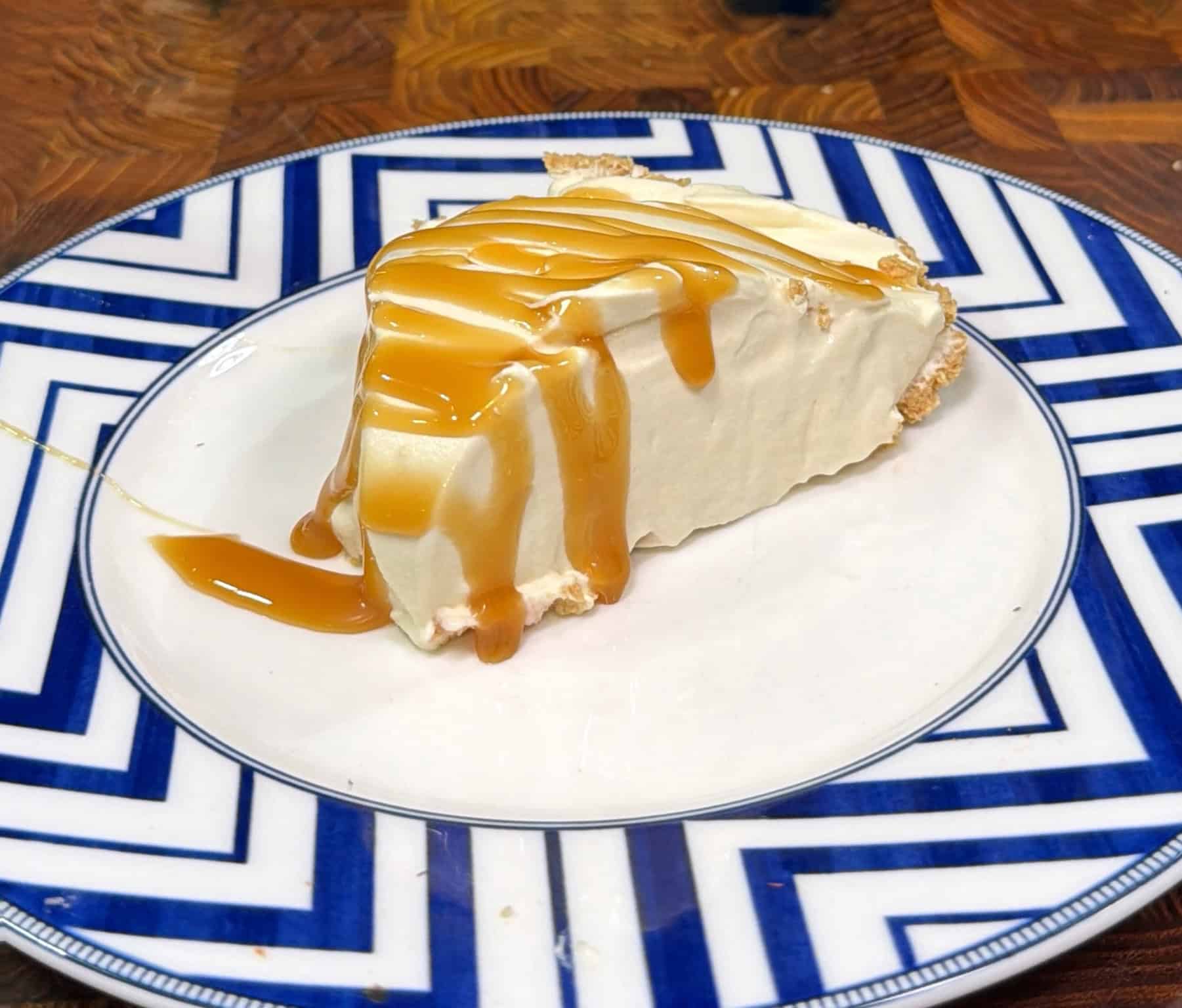 A slice of creamy cheesecake topped with caramel sauce sits on a white plate with a blue geometric pattern, placed on a wooden table.