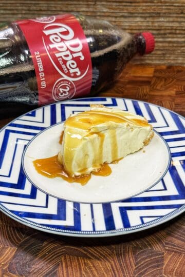 A slice of cheesecake drizzled with caramel sauce sits on a blue-and-white patterned plate, with a 2-liter bottle of Dr Pepper lying on its side in the background.