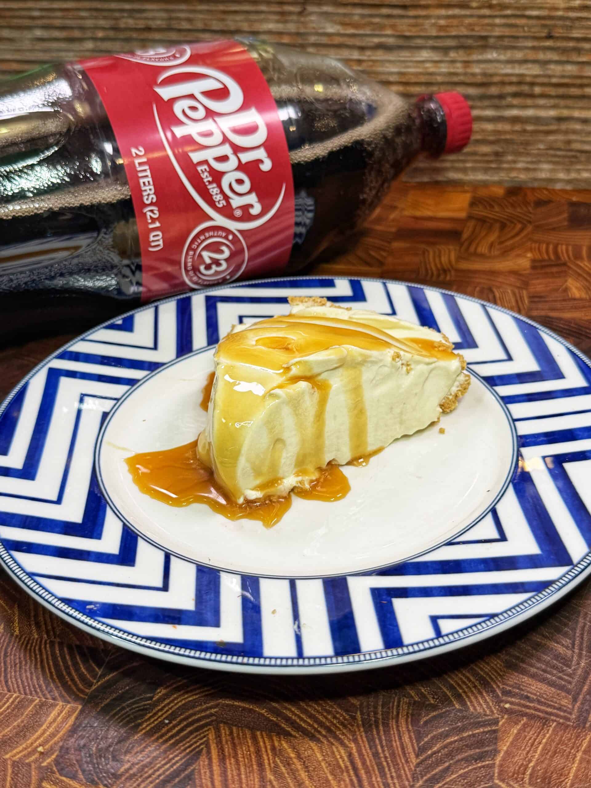 A slice of cheesecake drizzled with caramel sauce sits on a blue-and-white patterned plate, with a 2-liter bottle of Dr Pepper lying on its side in the background.