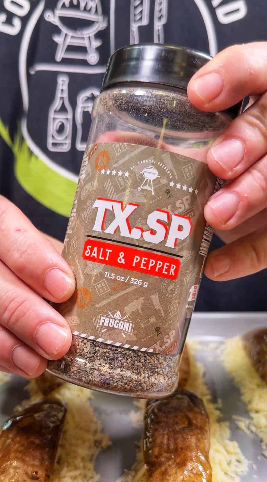 A person holds a container of Frugoni TX.SP Salt & Pepper seasoning over sausages covered with grated cheese, with a grill-themed shirt visible in the background.